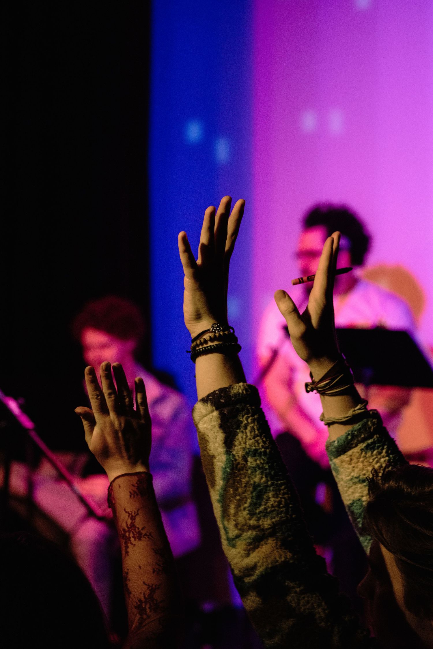 Raised hands with bracelets reach upward in front of a colorful purple and blue stage as musicians perform in the background.