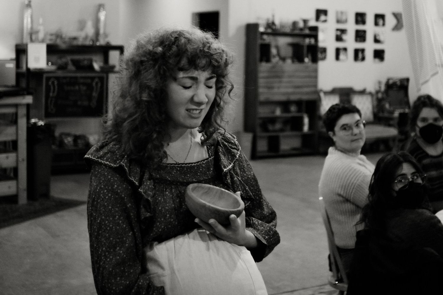 A curly-haired woman in a patterned cardigan holds a wooden bowl while seated audience members look on in a cozy, softly lit room.
