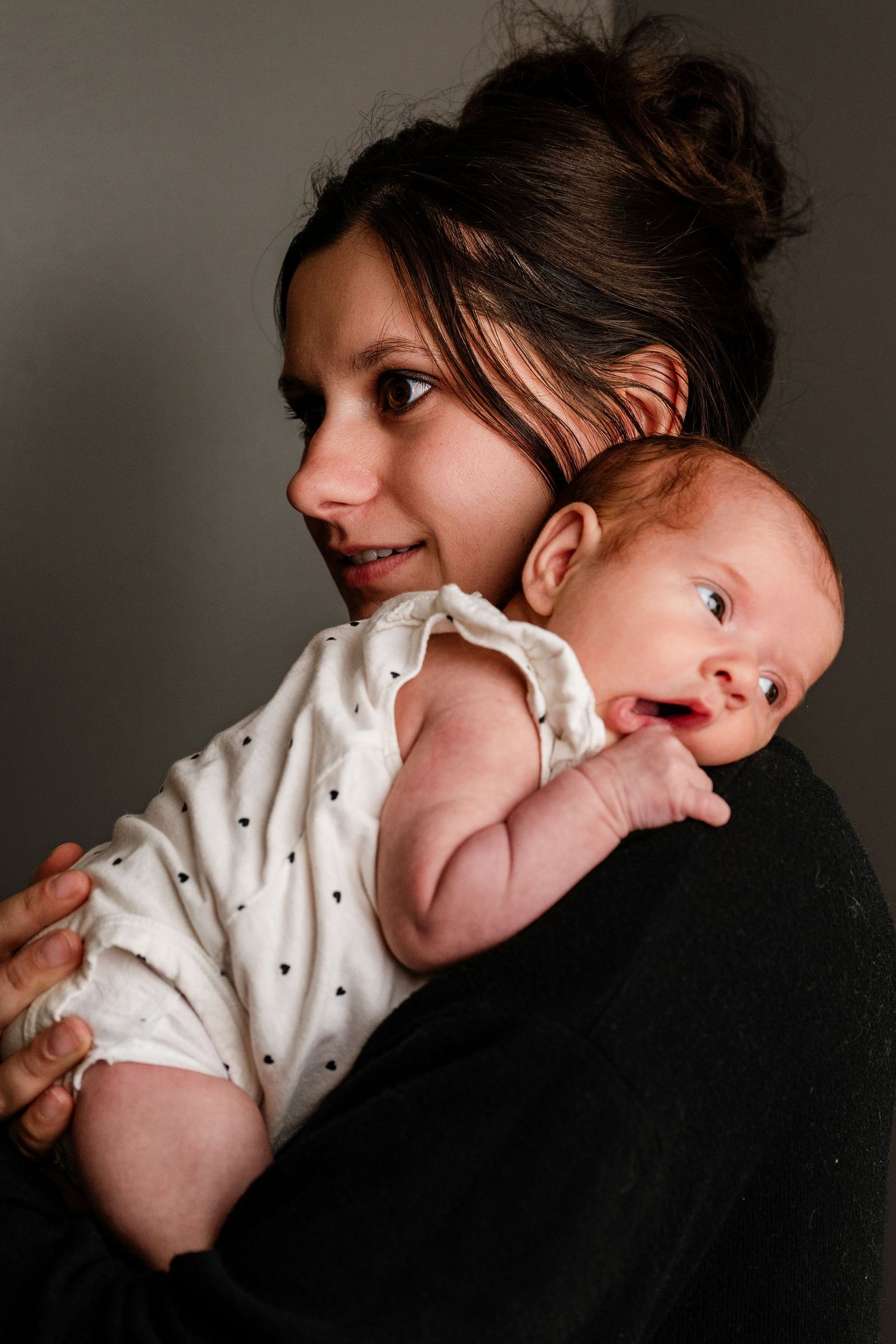 Mother with dark hair in a bun holds a newborn baby in a polka dot outfit against her shoulder.