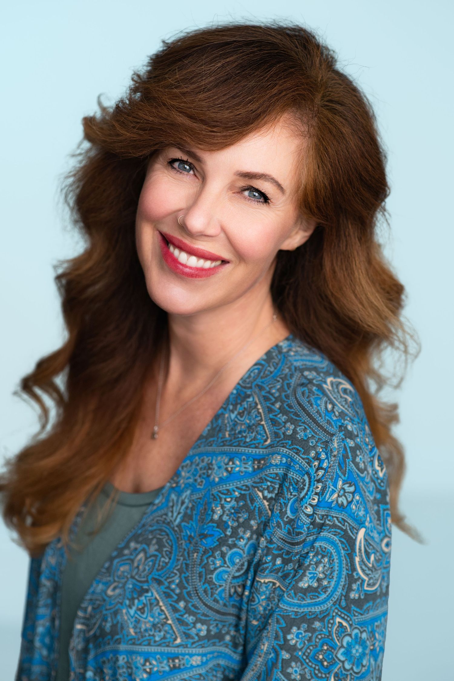 Smiling woman with long wavy auburn hair wears a blue paisley patterned top in a bright headshot against a soft pale blue background.