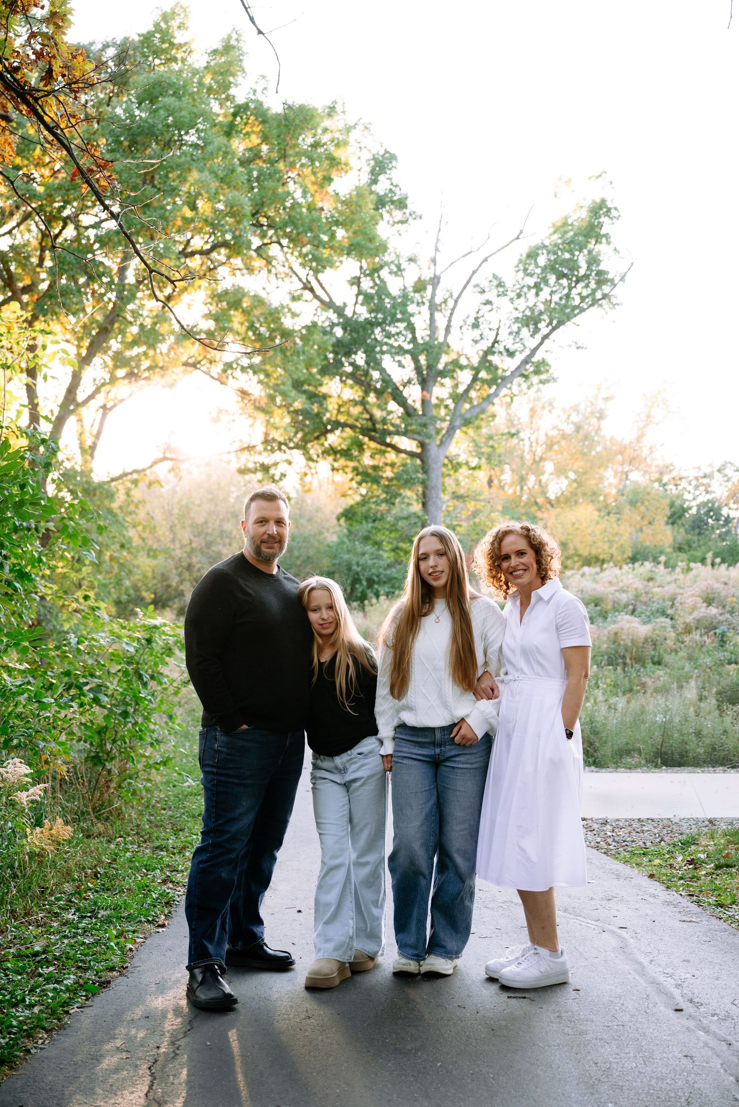 A family of four poses on a paved path in a park, with a man in black, two girls in jeans, and a woman in a white dress.