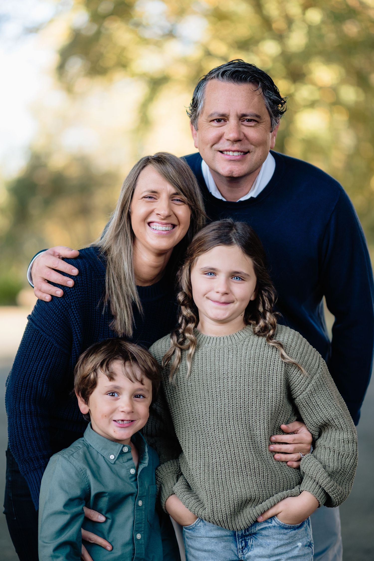 Smiling family of four in cozy sweaters pose together outdoors for a warm autumn portrait.