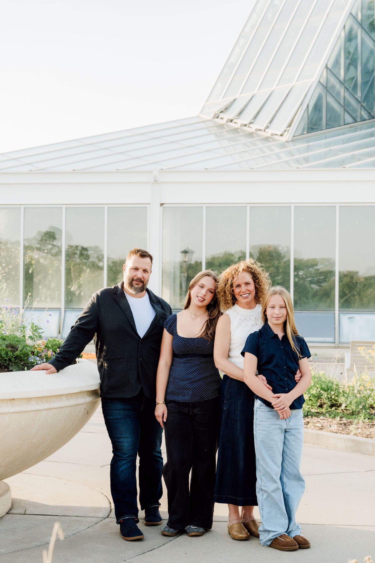 Family of four poses together outside a glass conservatory with greenery on a sunny day.