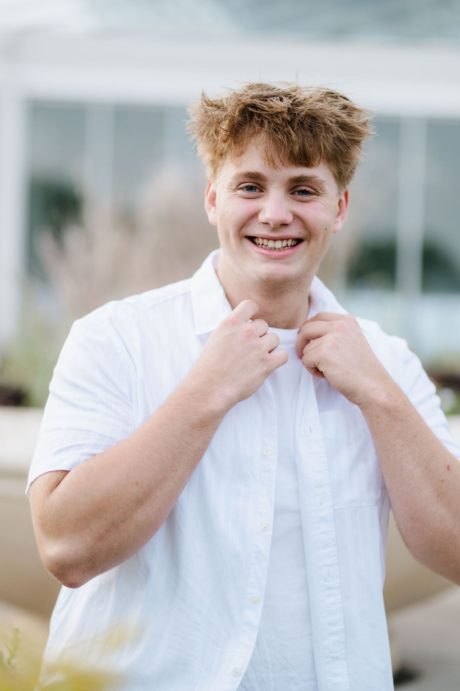 Smiling young man with tousled reddish-blond hair adjusts the collar of his white button-up shirt outdoors.
