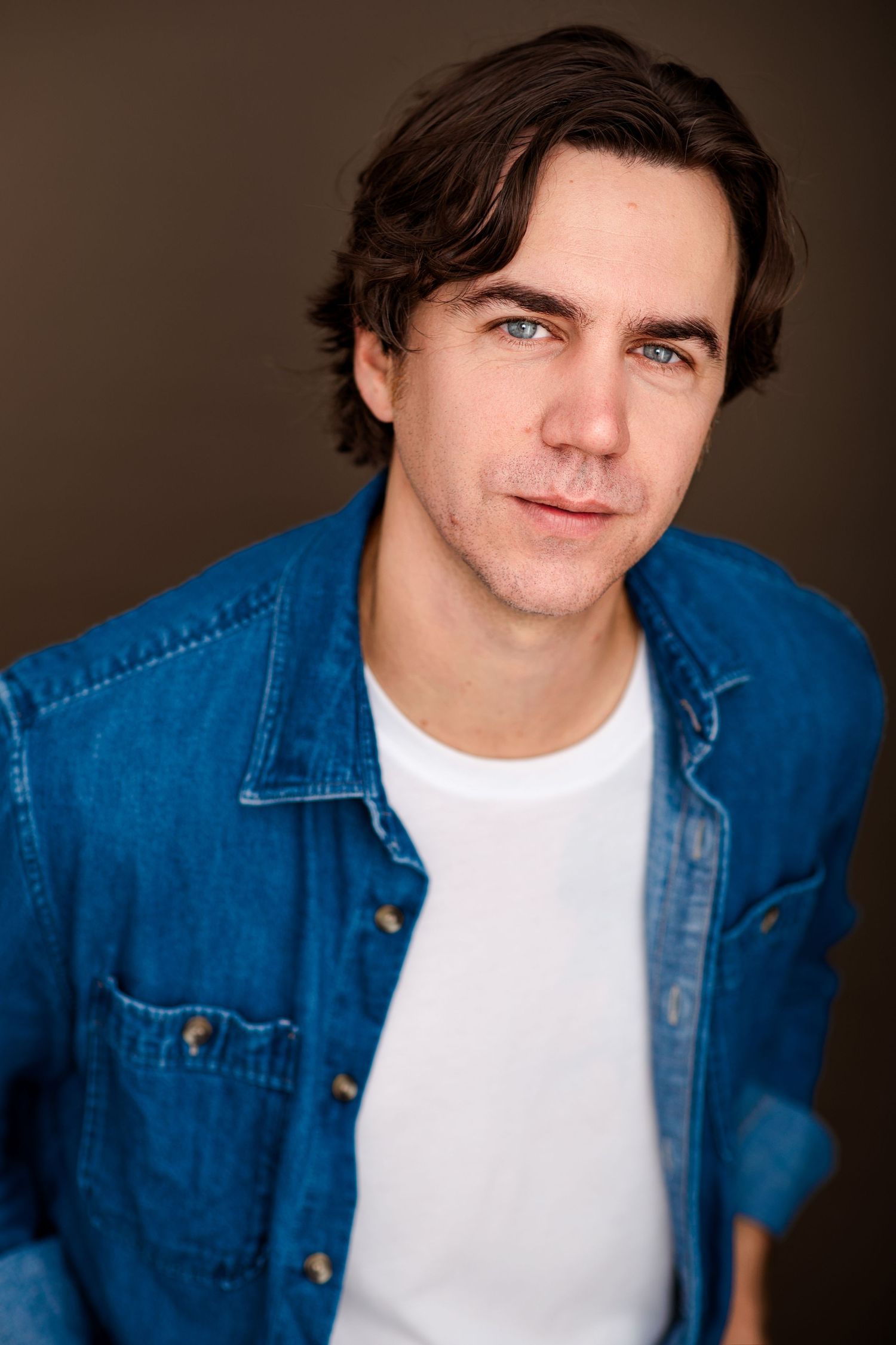 Headshot of a man with tousled dark brown hair and blue eyes wearing a denim jacket over a white t-shirt.
