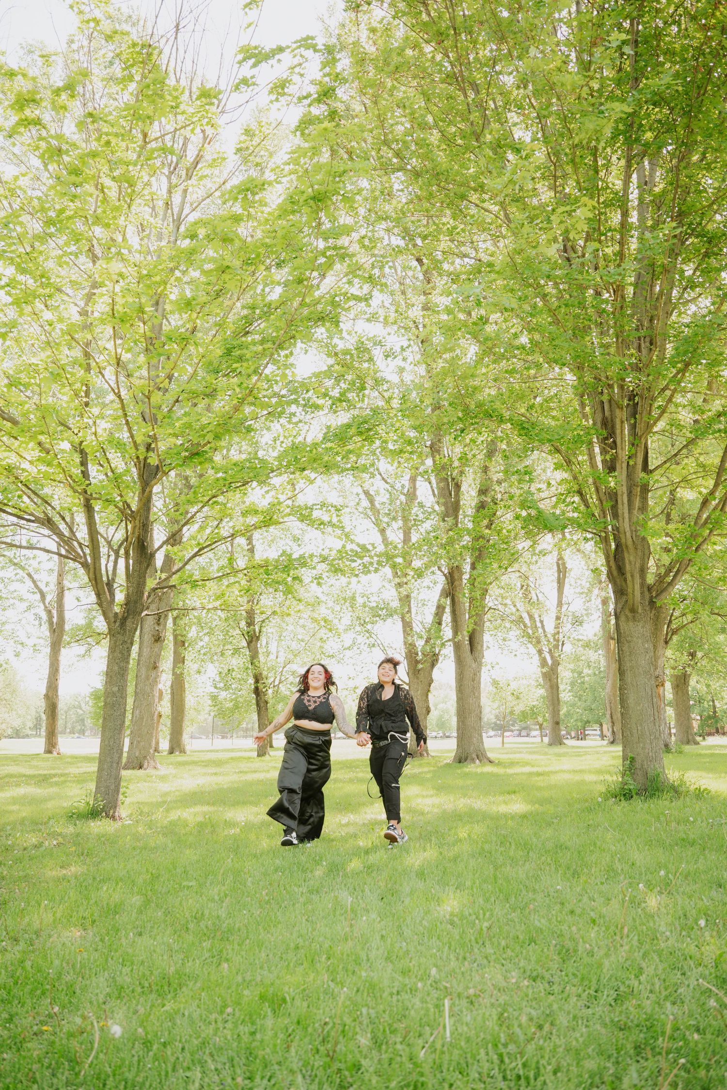 Two women in black outfits hold hands and run joyfully through a sunlit green park lined with tall leafy trees.
