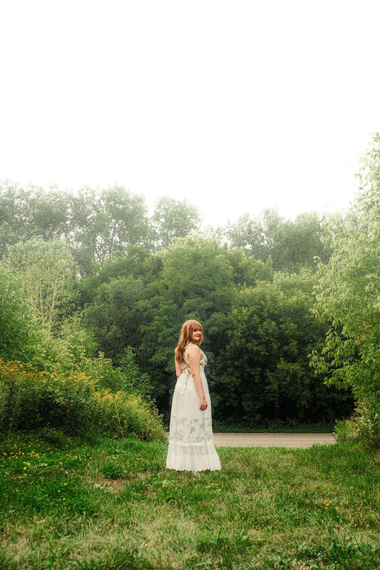 Woman in a long white floral dress looks over her shoulder while standing on a grassy lawn surrounded by lush green trees.
