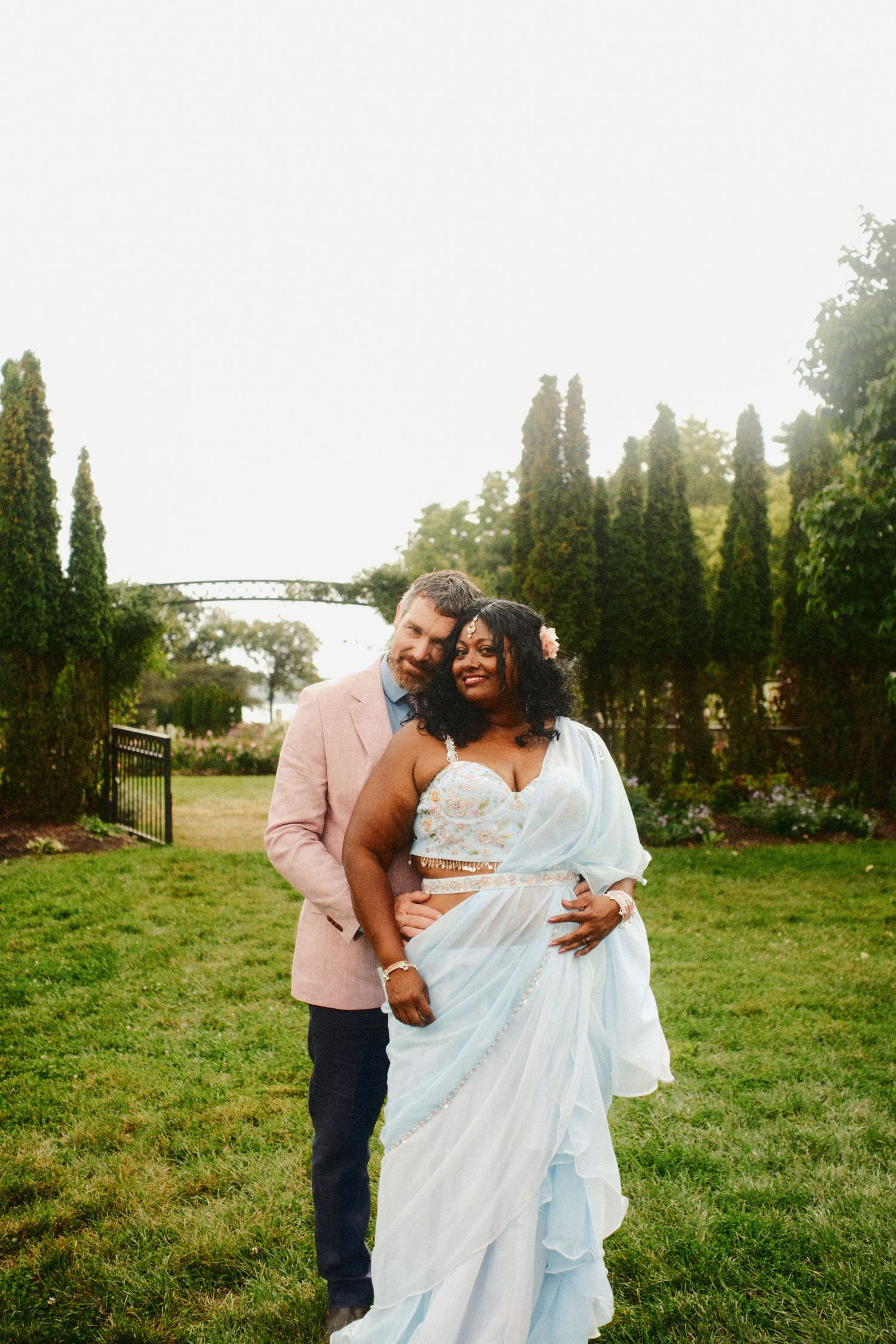 A smiling couple embraces on a green lawn, the bride in a flowing light blue sari and the groom in a blush pink blazer.