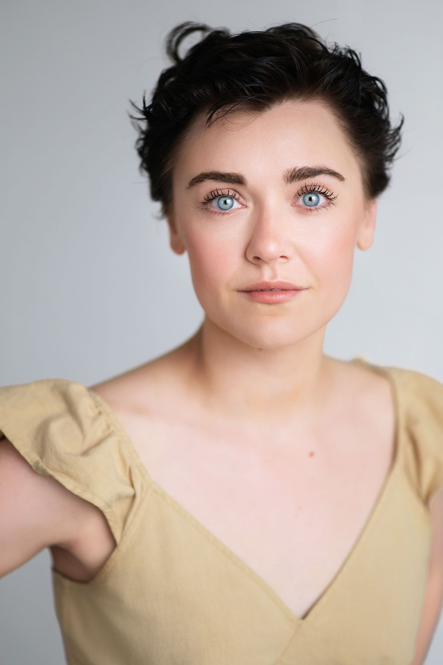 Woman with short dark pixie hair and bright blue eyes wears a tan top with ruffled sleeves in a studio portrait on a gray backdrop.