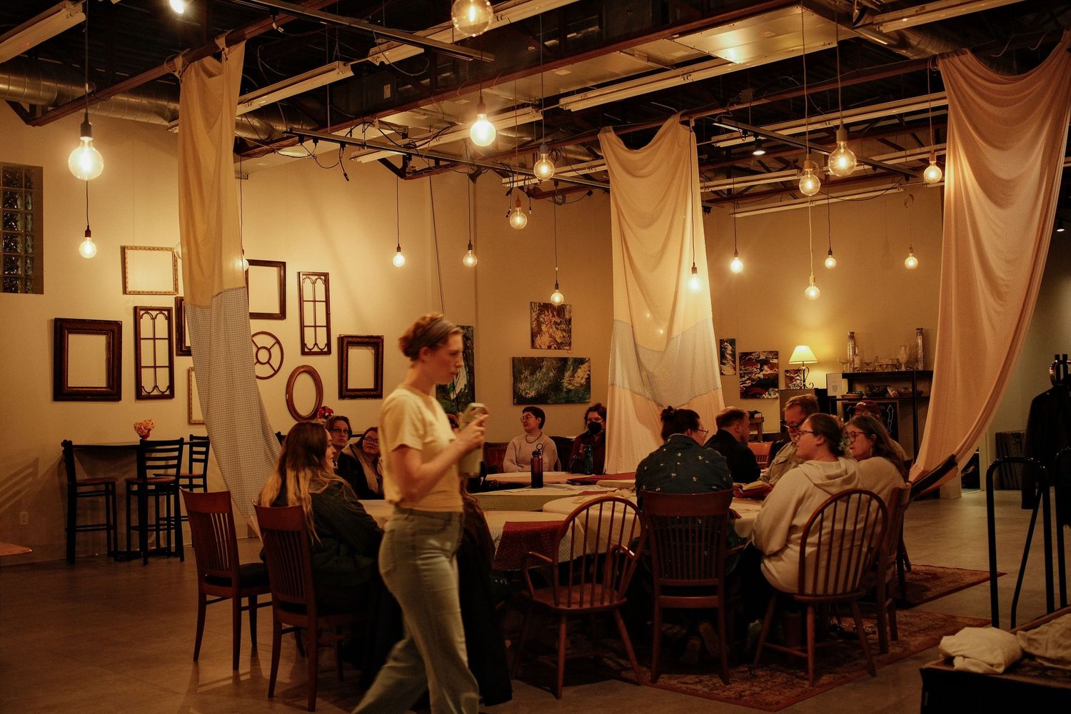 A woman stands speaking to guests seated around a long table in a warm, rustic venue with draped fabric and hanging string lights.