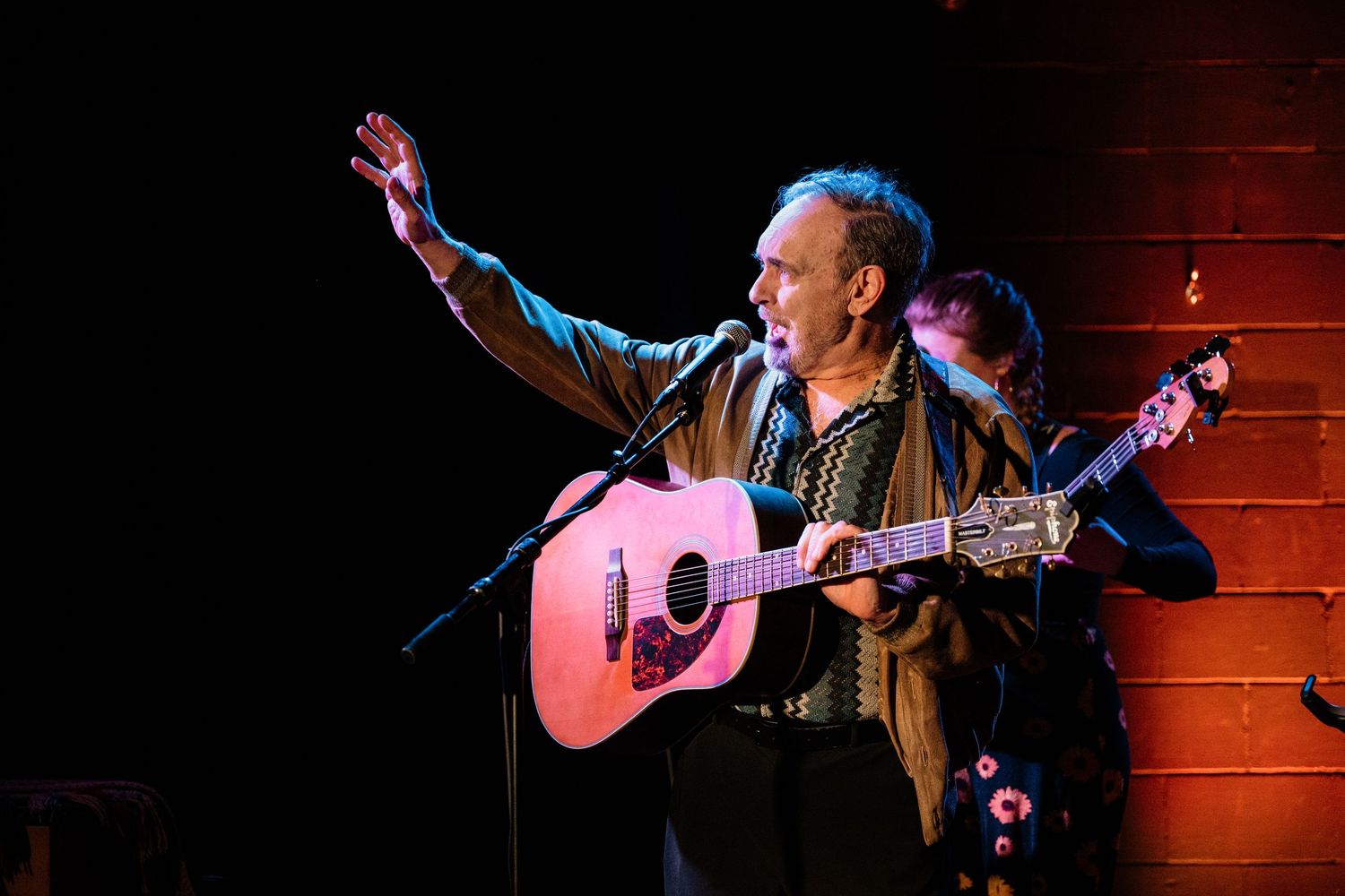 A man in a brown jacket plays an acoustic guitar and raises one arm while singing into a microphone on a dimly lit stage.