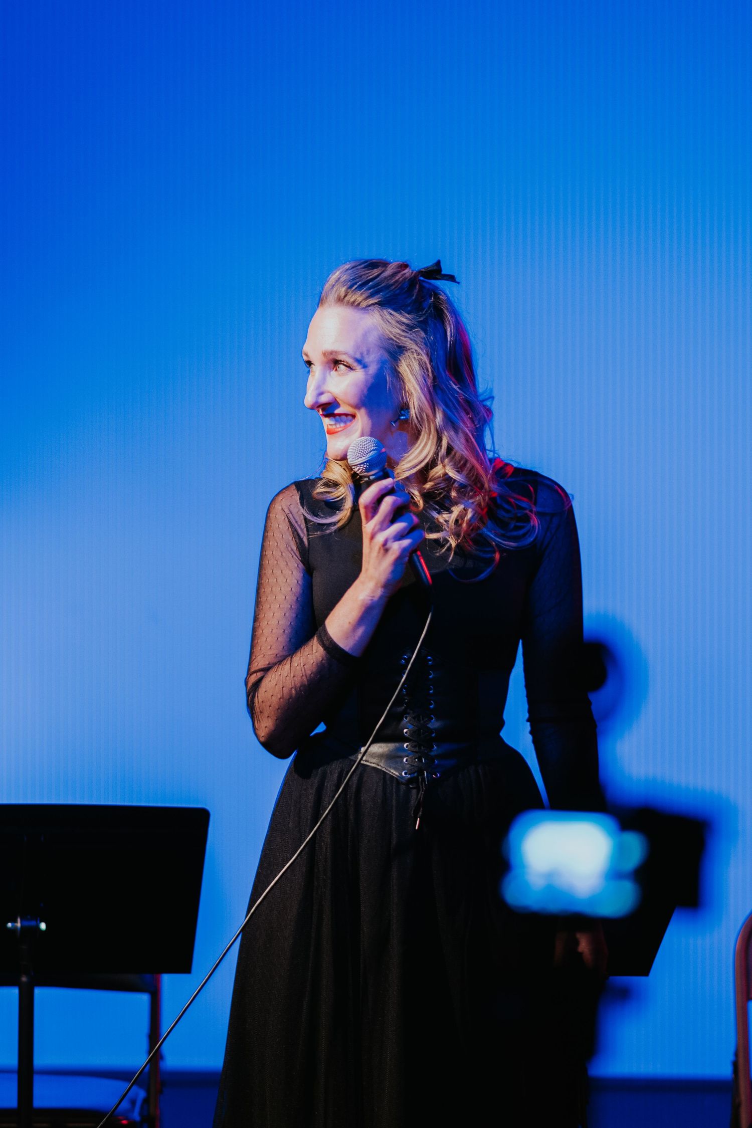 Smiling woman with wavy blonde hair holds a microphone on stage, wearing a black sheer-sleeved dress against a bright blue backdrop.
