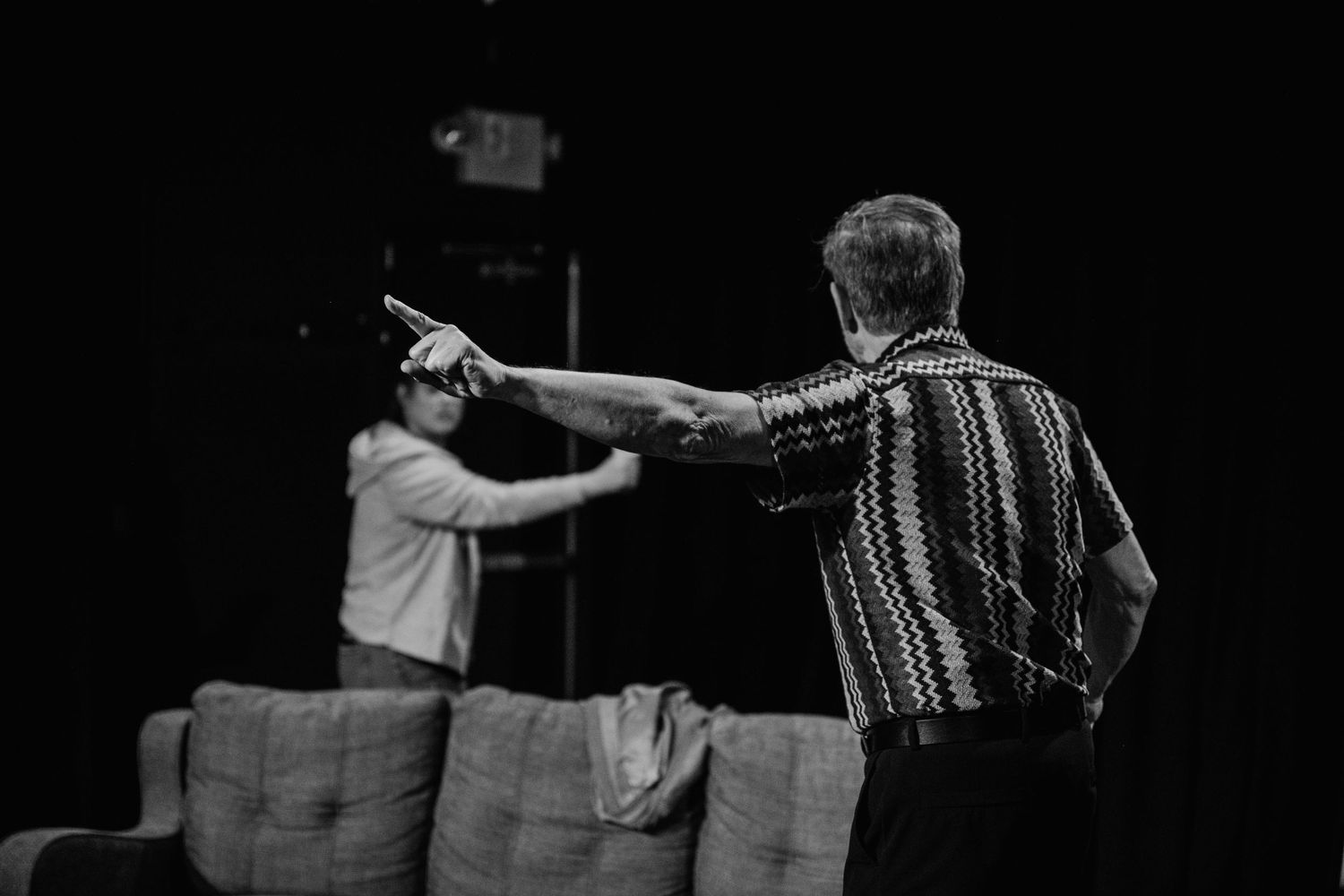 In a black and white stage scene, a man in a patterned shirt points across a couch toward another performer.