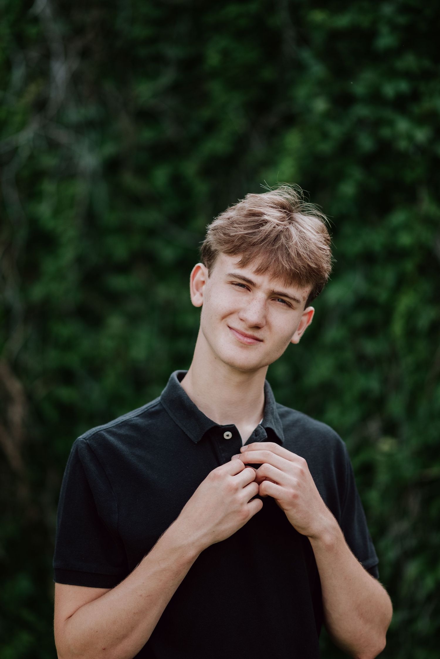 Smiling young man in a black polo shirt adjusts his collar in front of a lush green ivy wall.