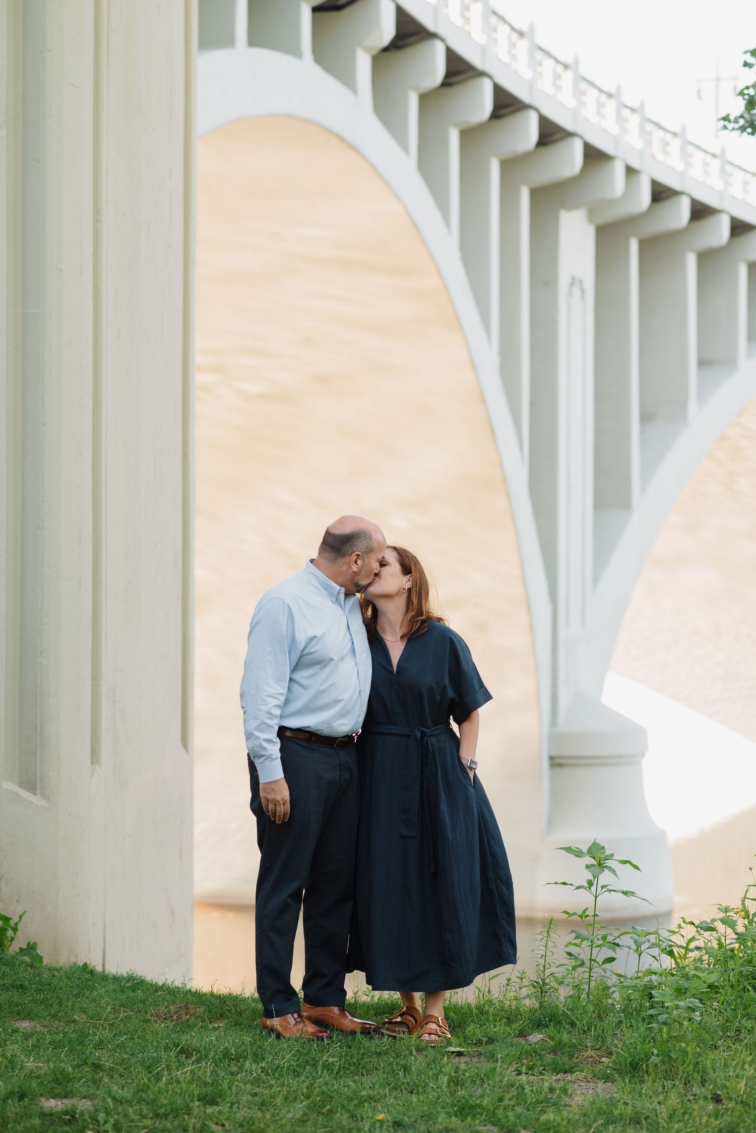 A couple shares a kiss on a grassy hillside beneath a large white arched stone bridge during an engagement photo session.
