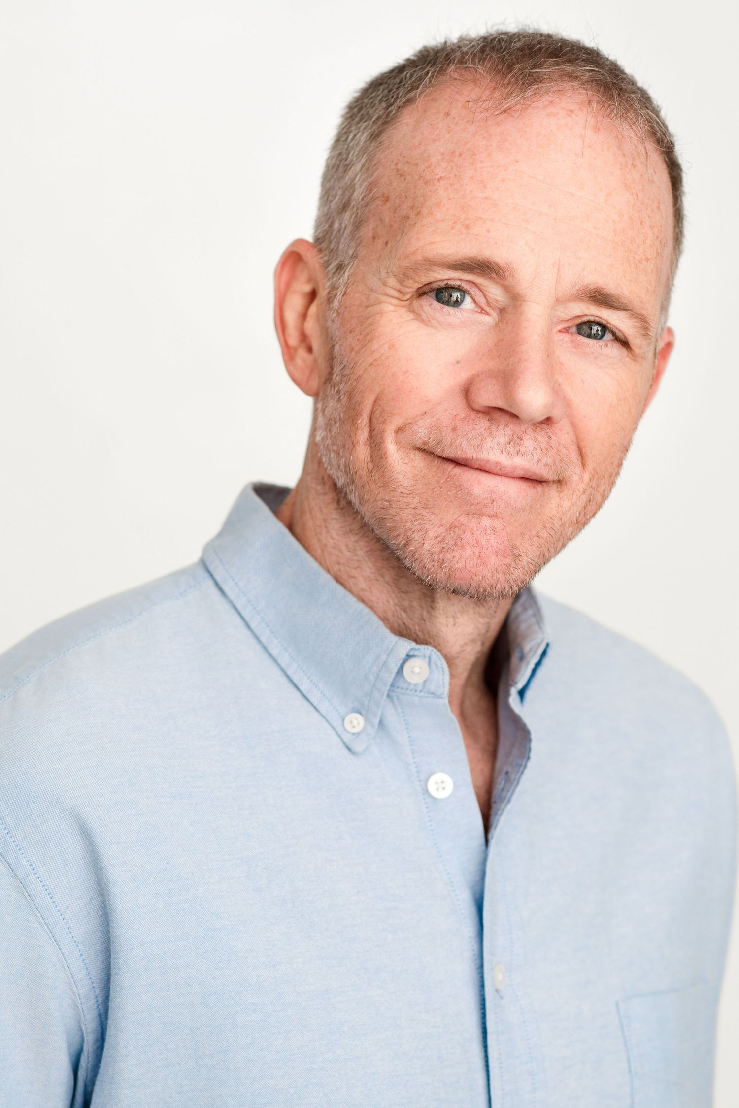 Headshot of a smiling middle-aged man with short gray hair and light stubble wearing a light blue button-down shirt.