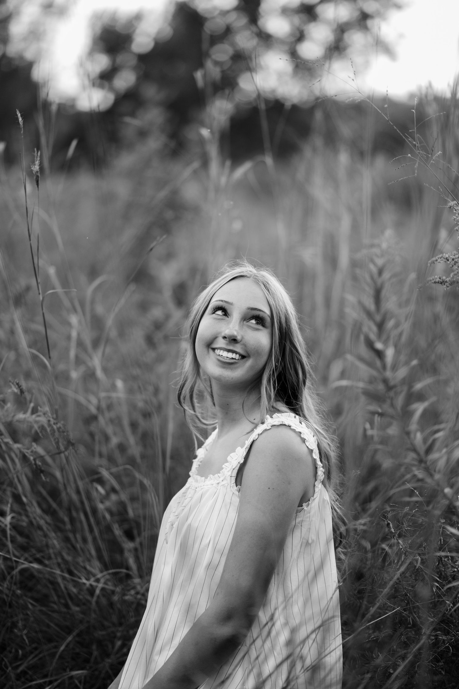 Black and white portrait of a smiling young woman in a lace-trimmed sundress looking upward amid tall grasses in a field.
