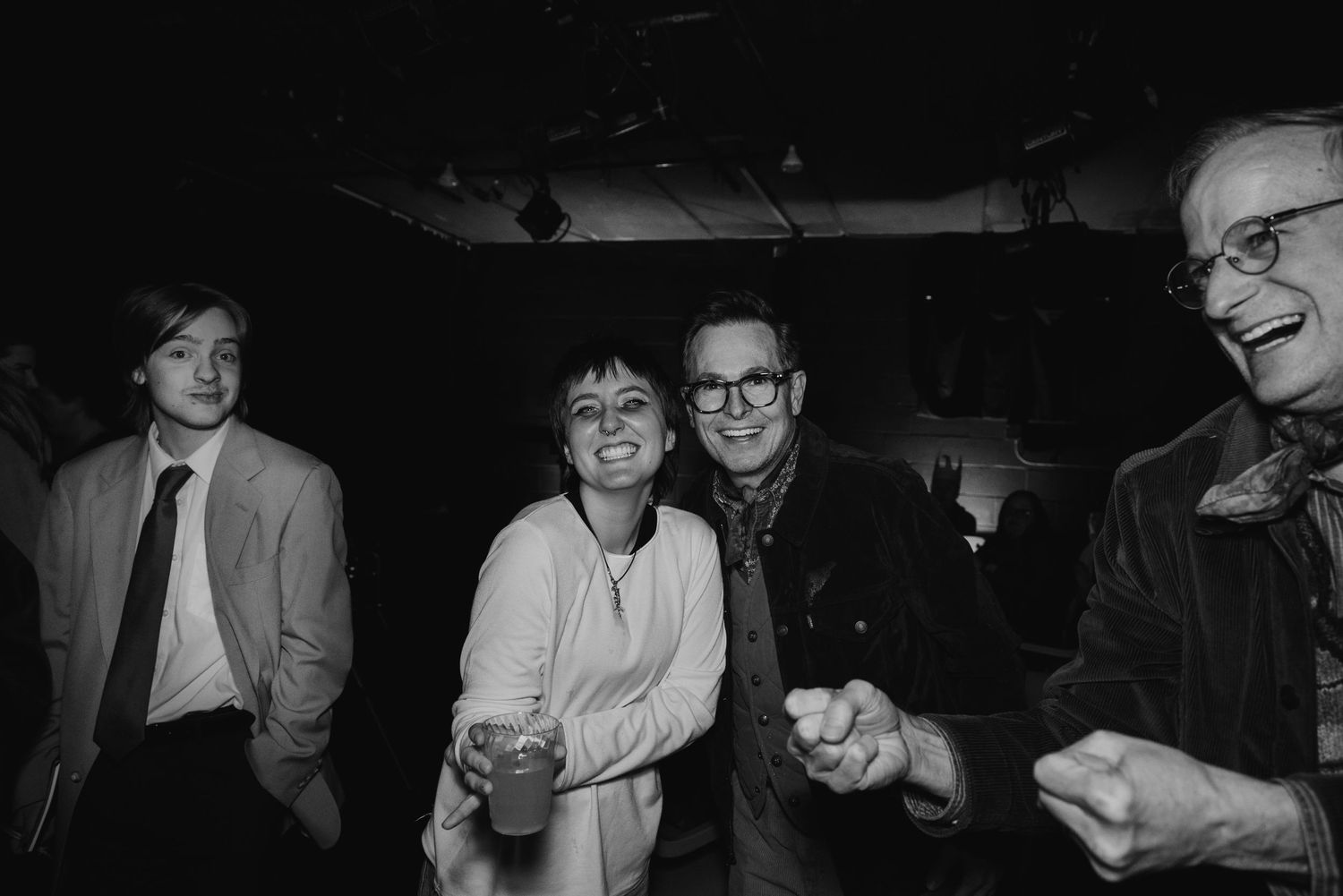 Four smiling people in a dimly lit room laugh and dance together at a lively indoor party, one holding a drink.
