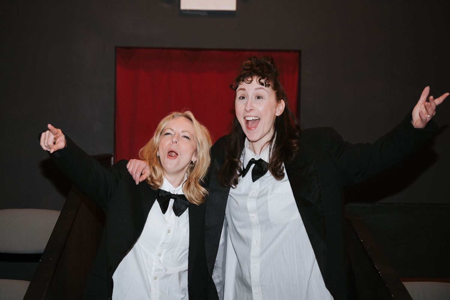 Two laughing performers in black blazers, white shirts and bow ties pose with arms raised in front of a red curtain on stage.