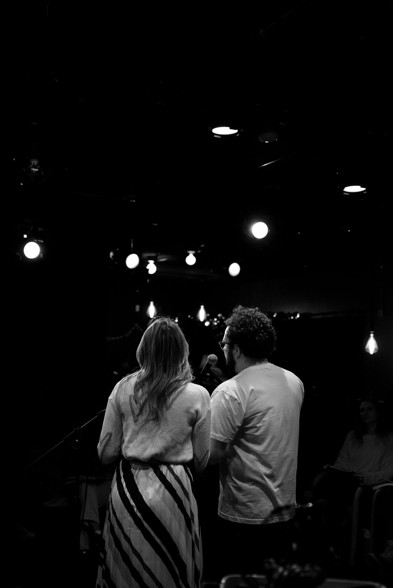 Black and white photo of a woman and a man seen from behind facing a stage with glowing string lights above.