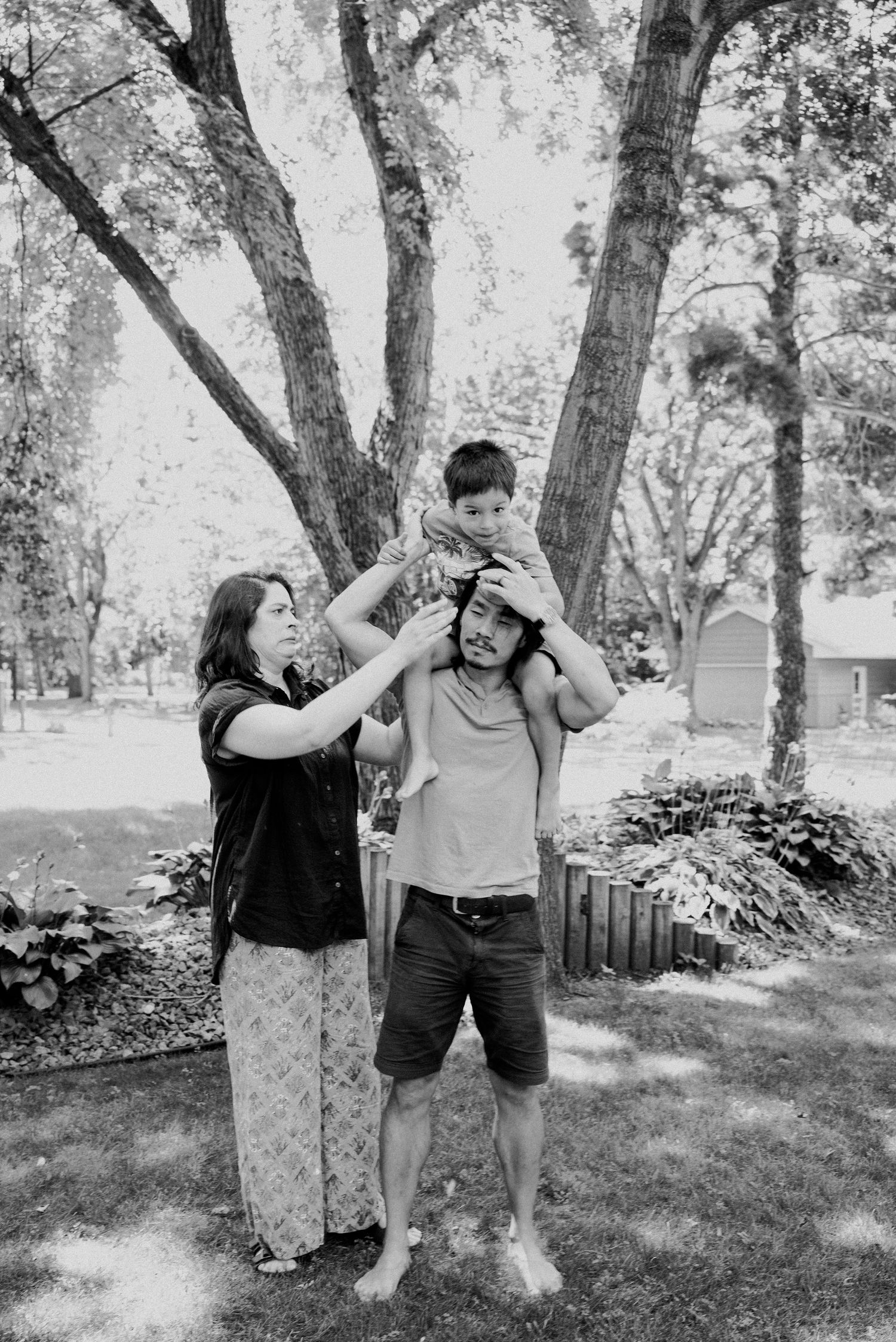 A black and white photo of a woman reaching up to a young boy sitting on a barefoot man's shoulders beneath a large tree.