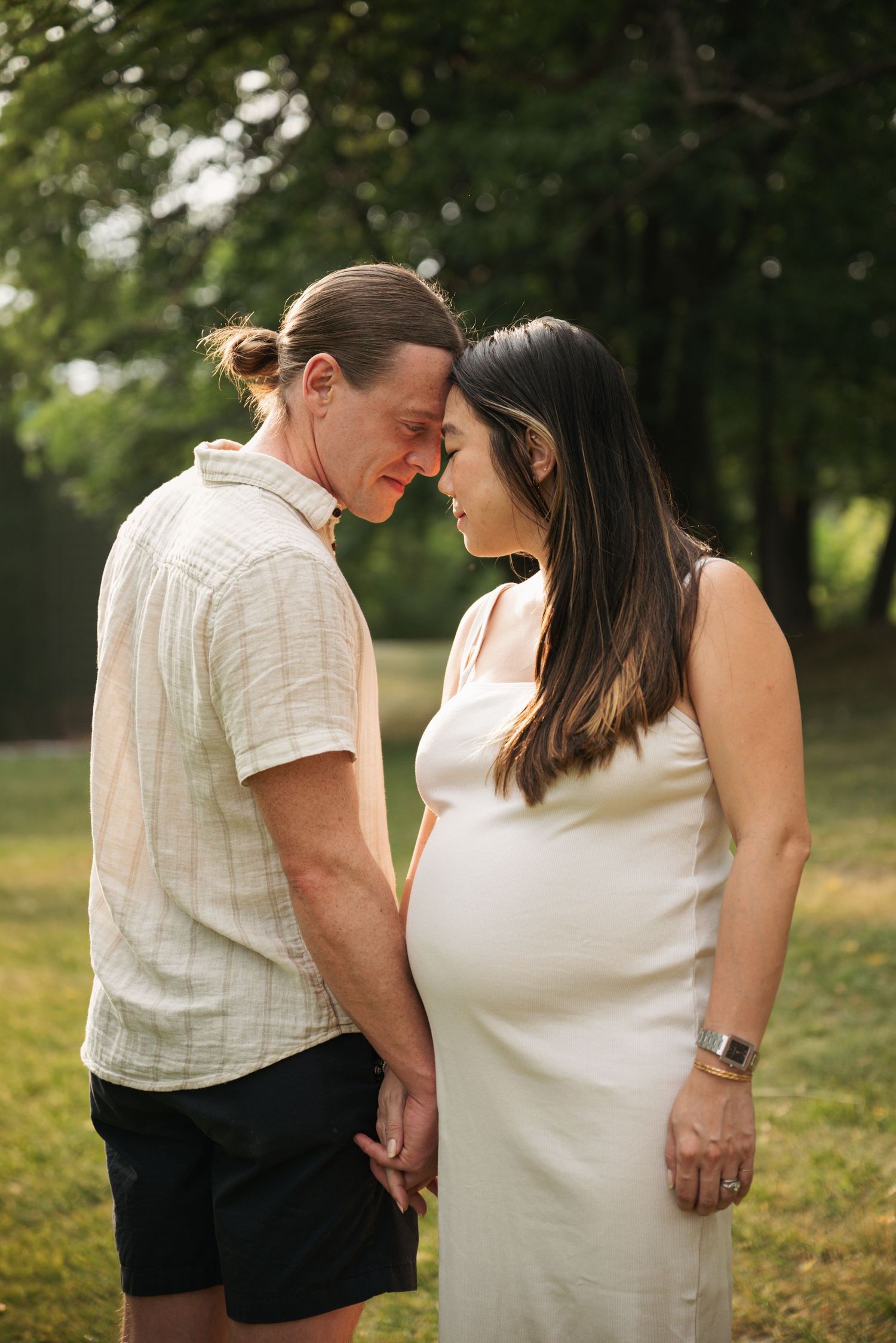 A man and a pregnant woman in a cream dress touch foreheads and hold hands in a sunlit park during a maternity photo session.