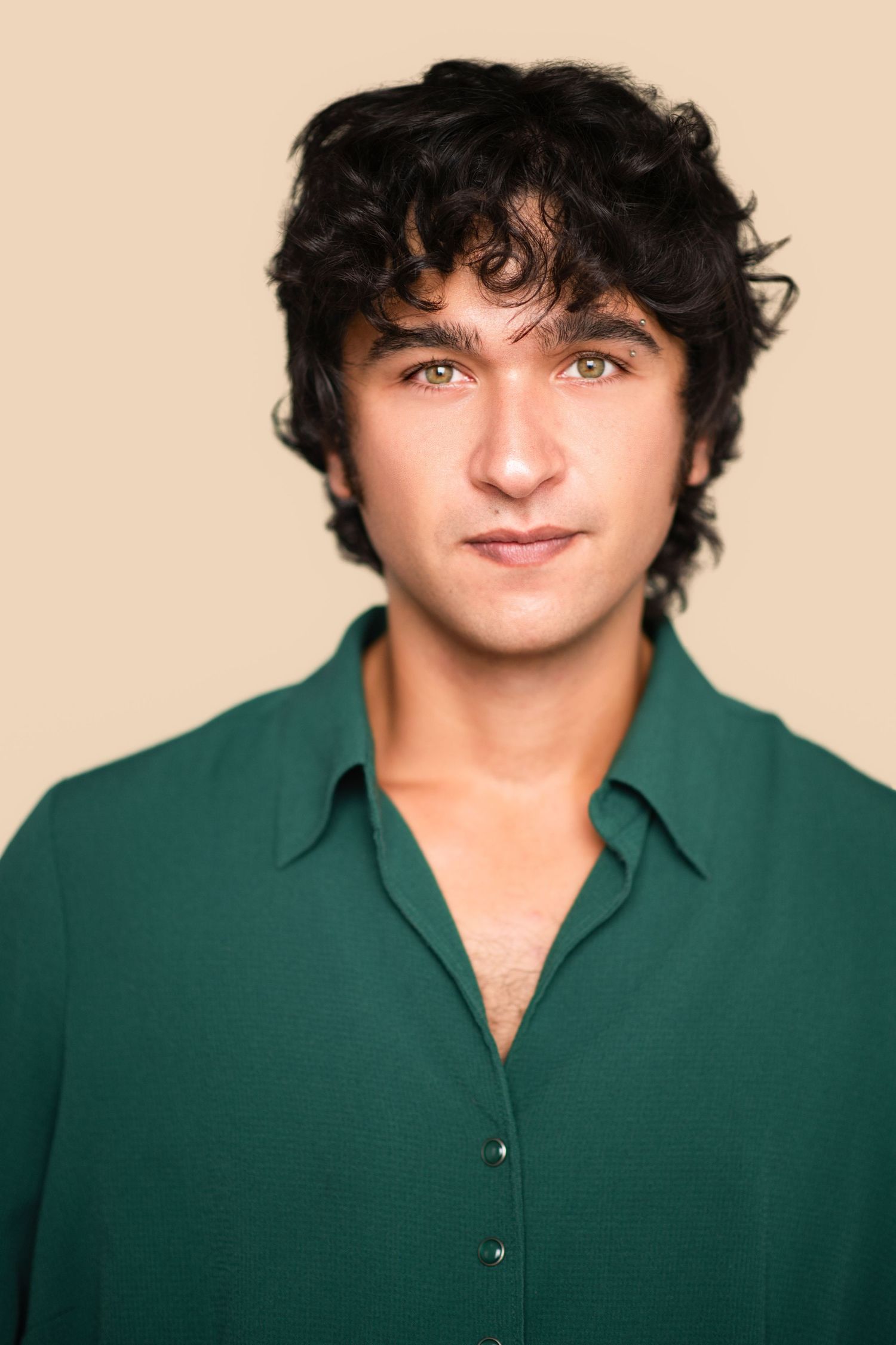 Young man with dark curly hair wearing an emerald green button-up shirt poses against a neutral beige studio backdrop.