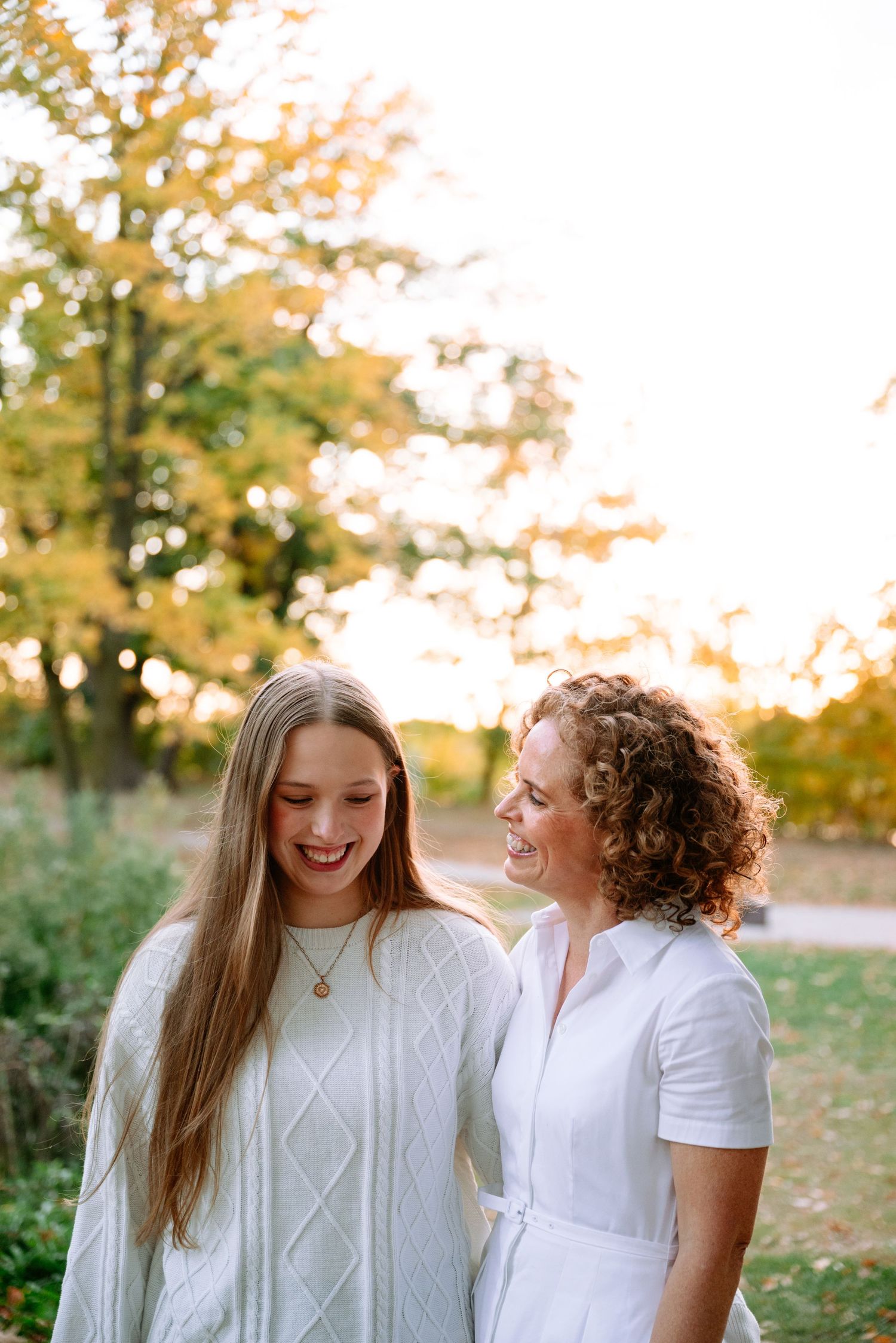 A smiling teenage girl with long hair in a white knit sweater stands beside a laughing curly-haired woman in a white dress outdoors.