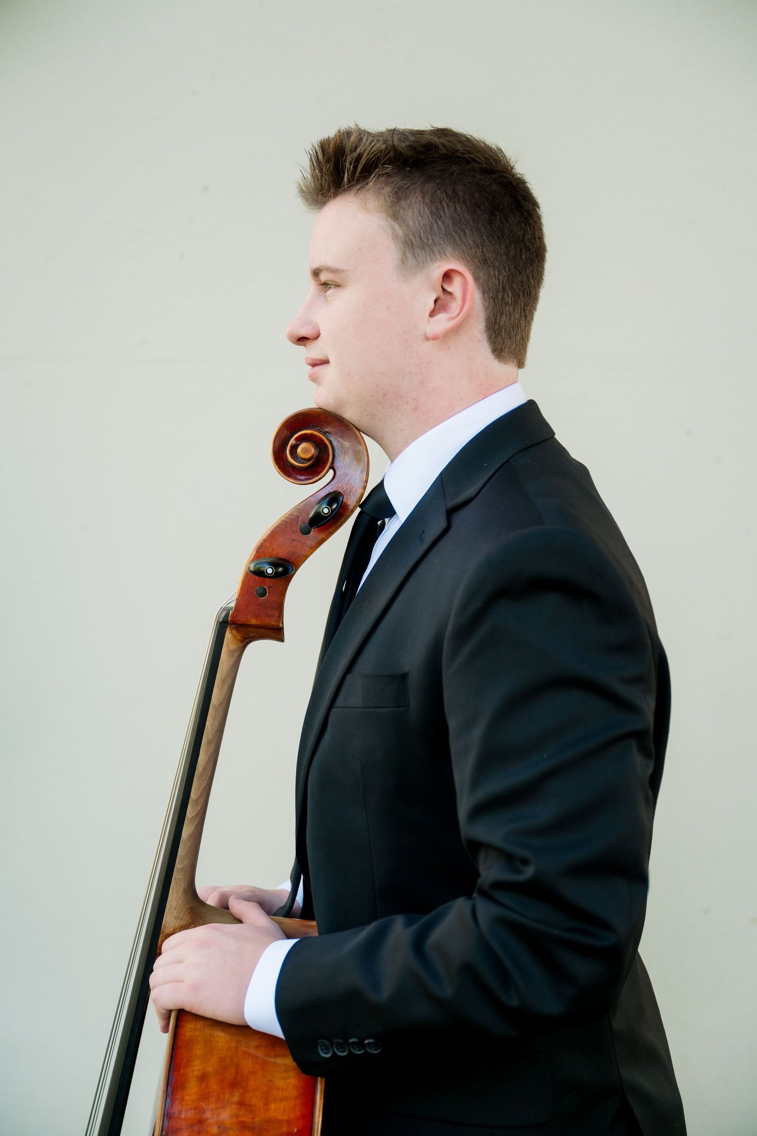 Young man in a black suit and tie stands in profile holding a cello against a plain light wall in a classical musician portrait.
