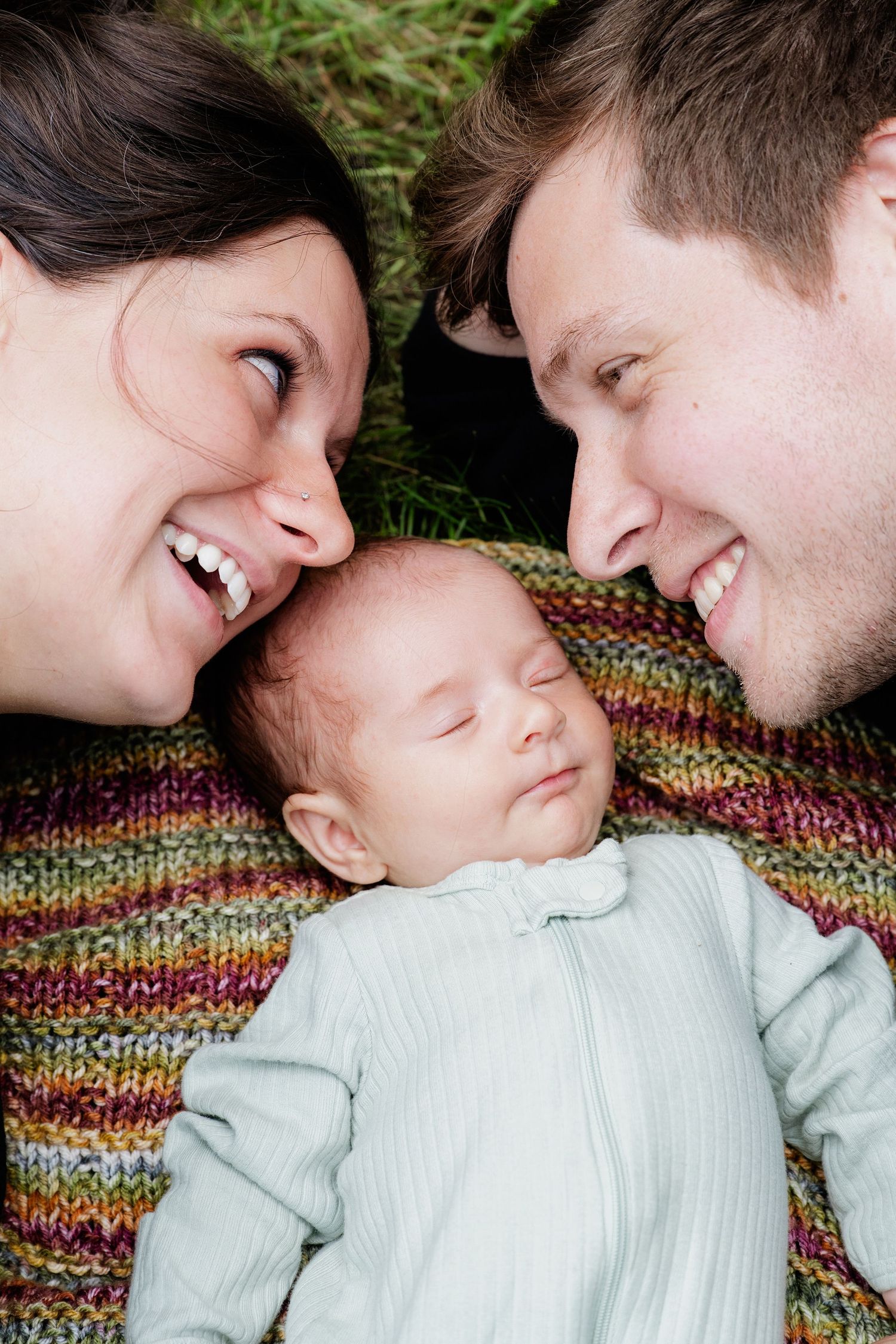 Smiling parents lie on a colorful striped blanket in the grass, gazing lovingly at their sleeping newborn baby between them.