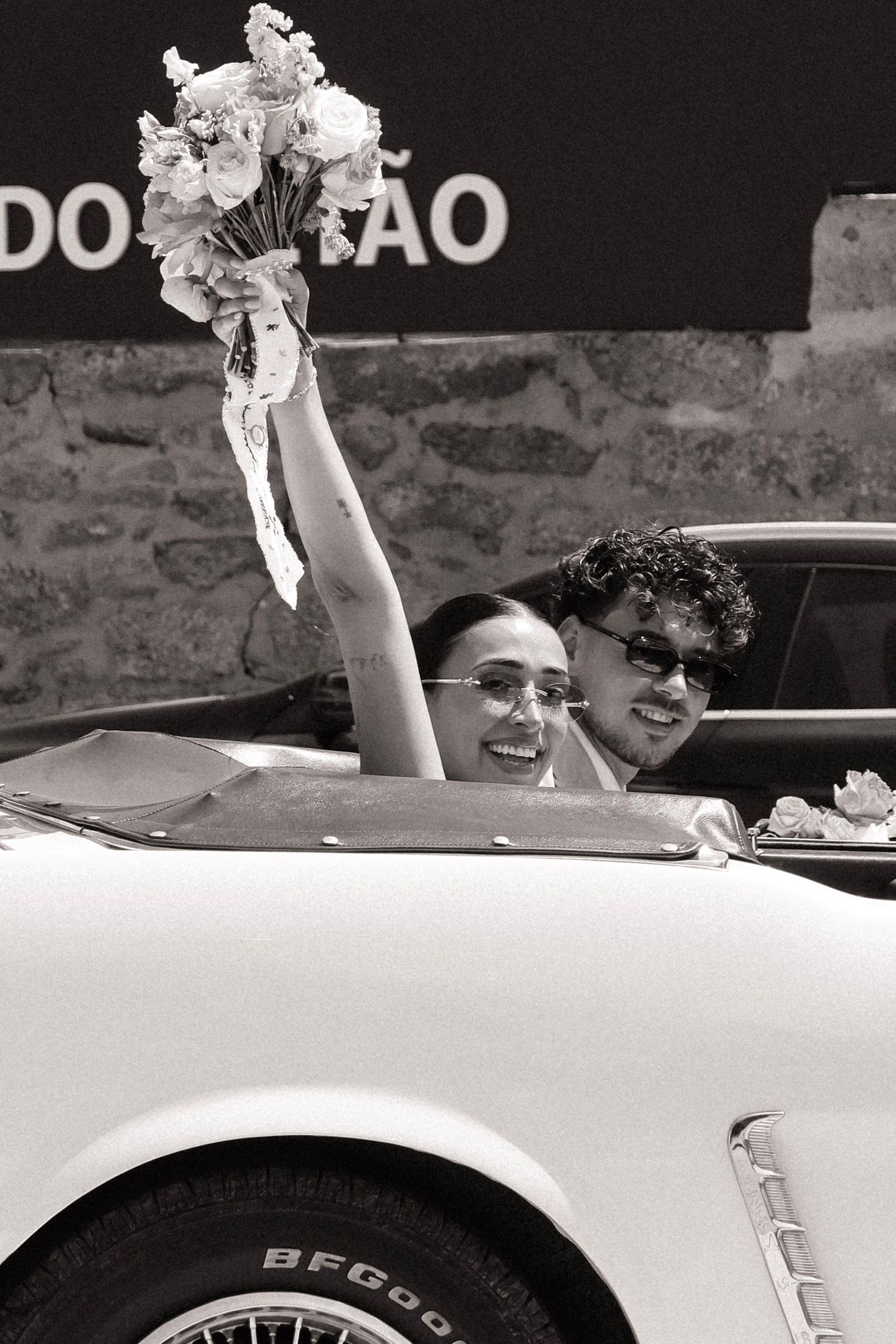 Joyful newlyweds in vintage white convertible car, bride raising flower bouquet, black and white wedding photo.