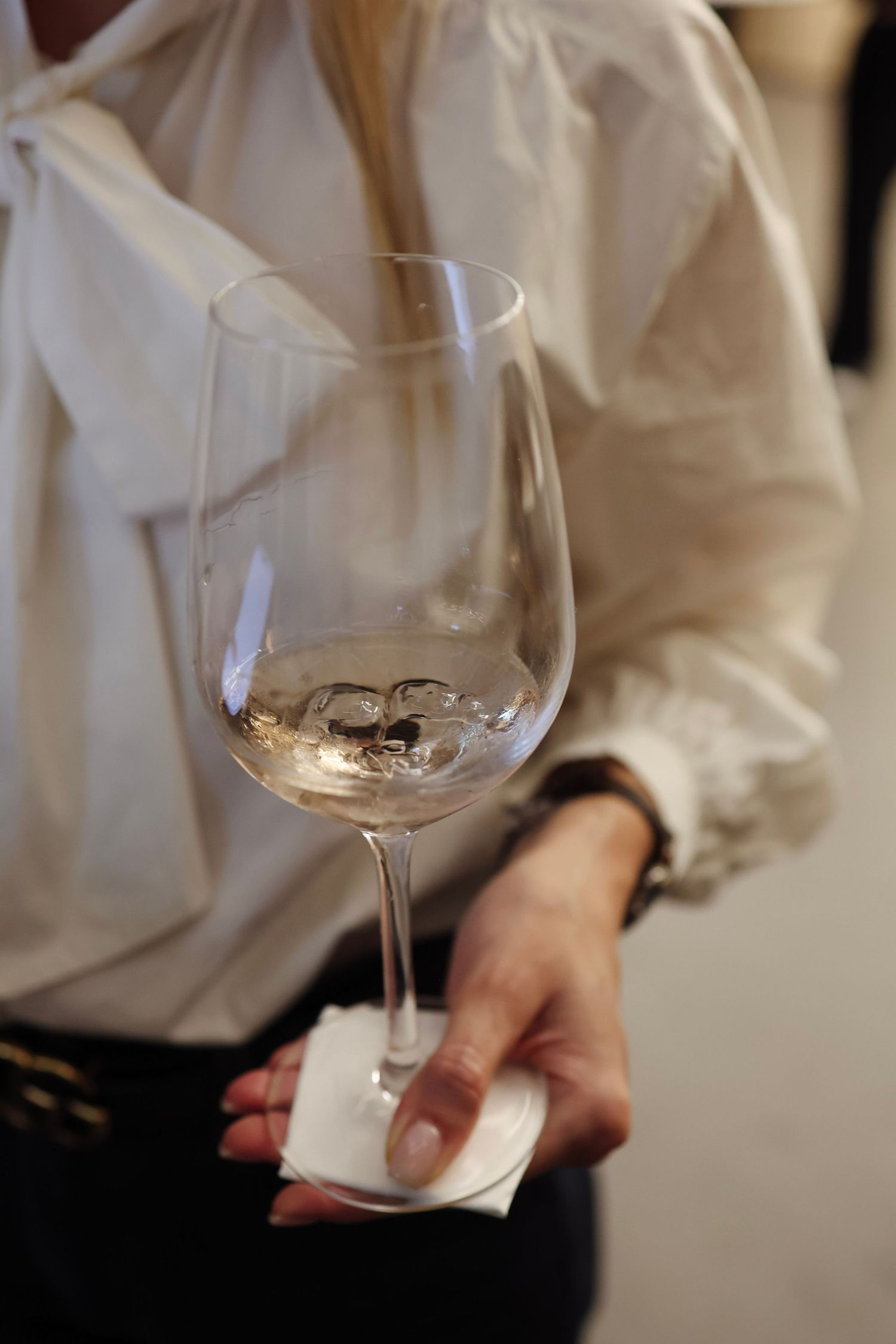 Elegant woman in white ruffled blouse holding a crystal wine glass with white wine at an event.