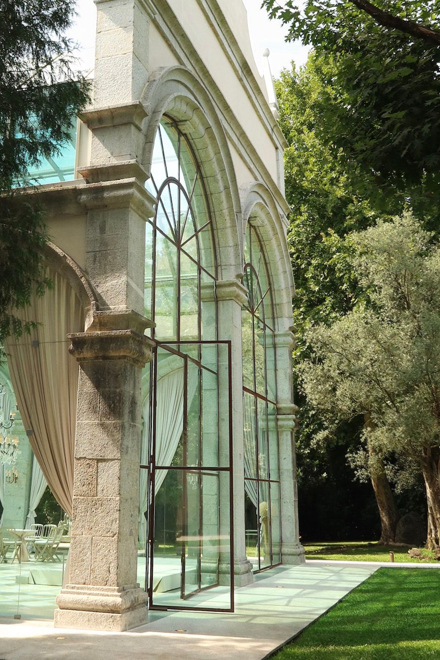 Elegant wedding venue with gothic stone arches and modern glass walls surrounded by lush garden trees.