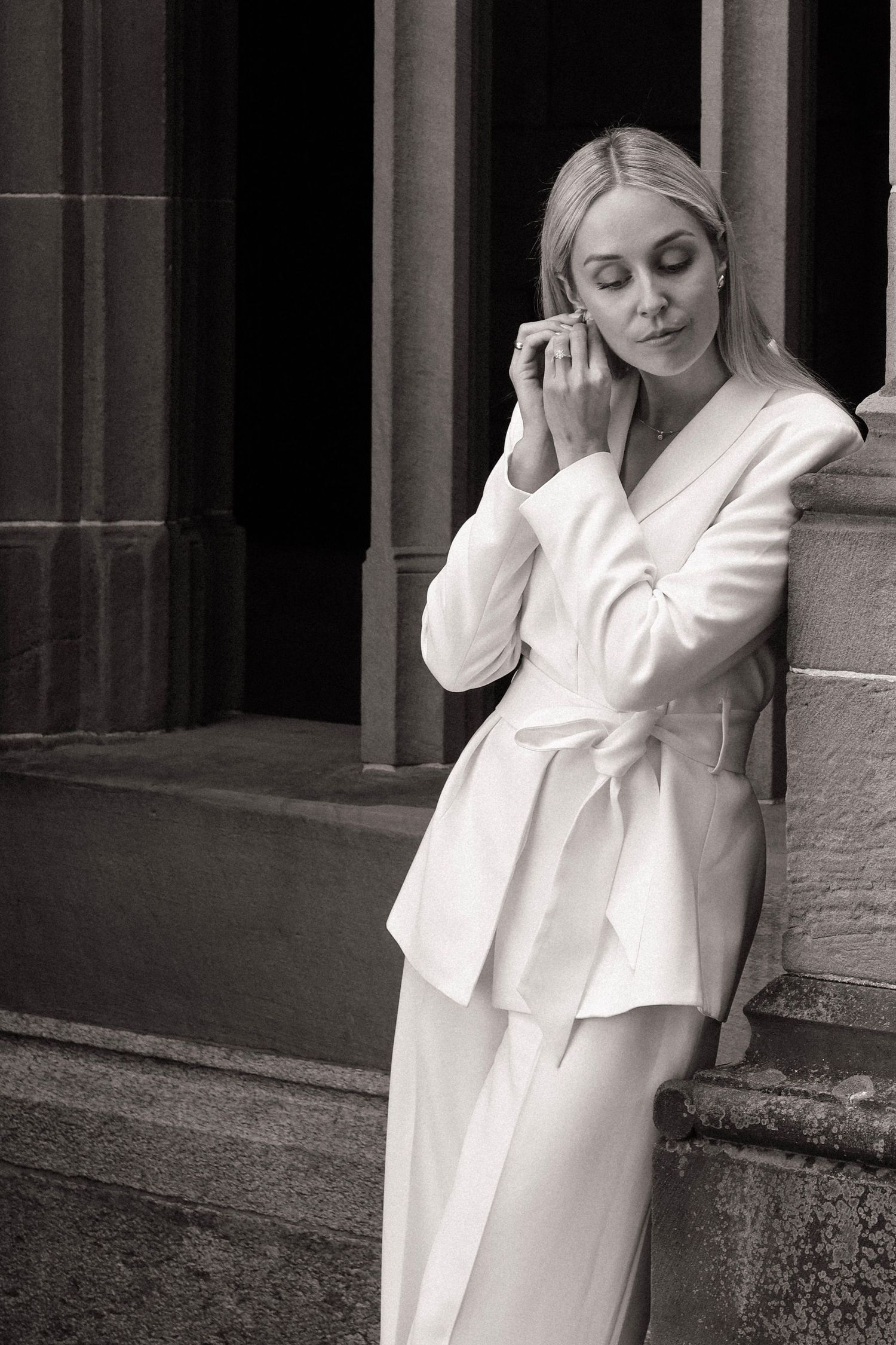 Elegant bride in white belted blazer pantsuit leaning against stone column in black and white.