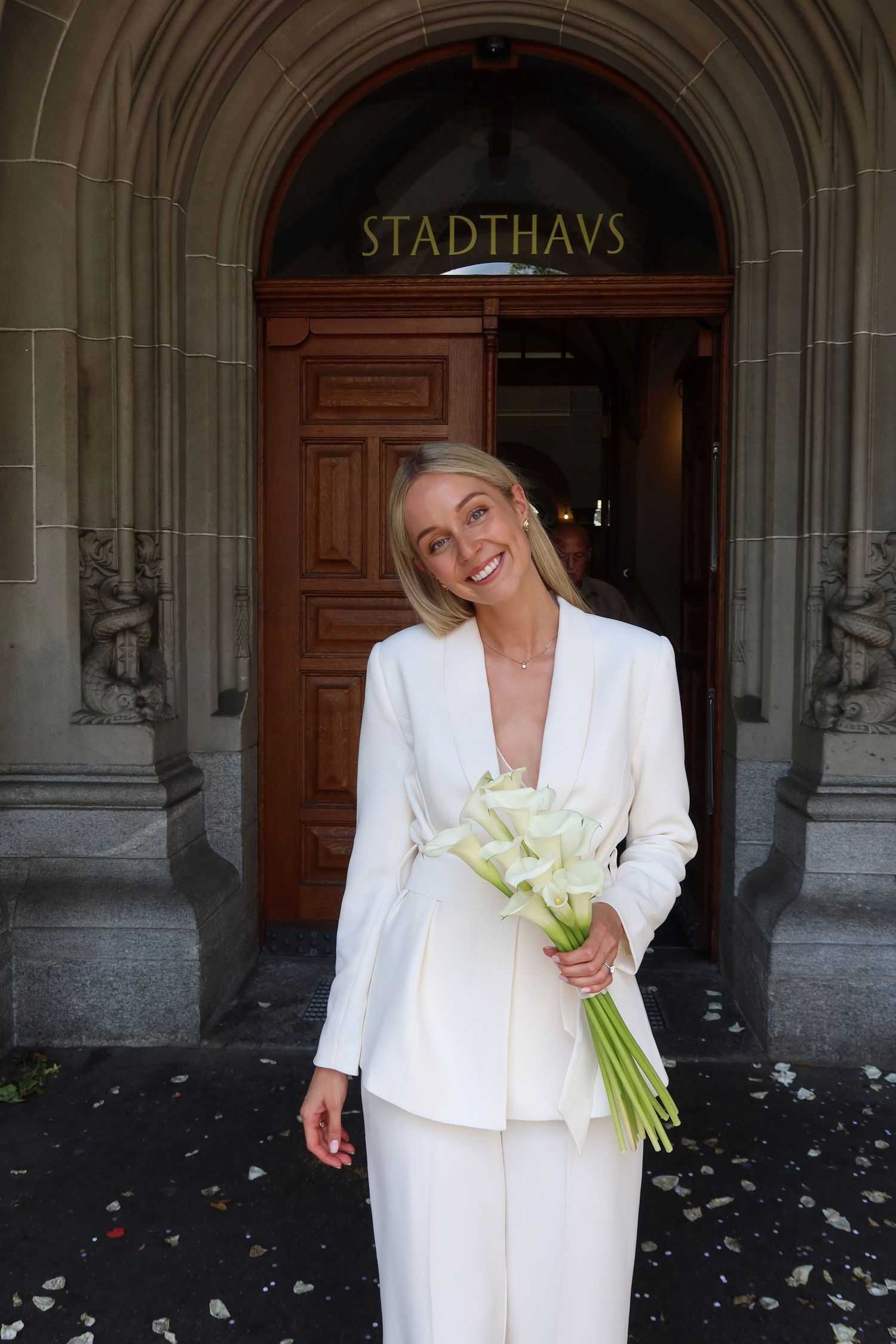 Smiling blonde bride in white pantsuit holding calla lily bouquet outside Stadthaus entrance.