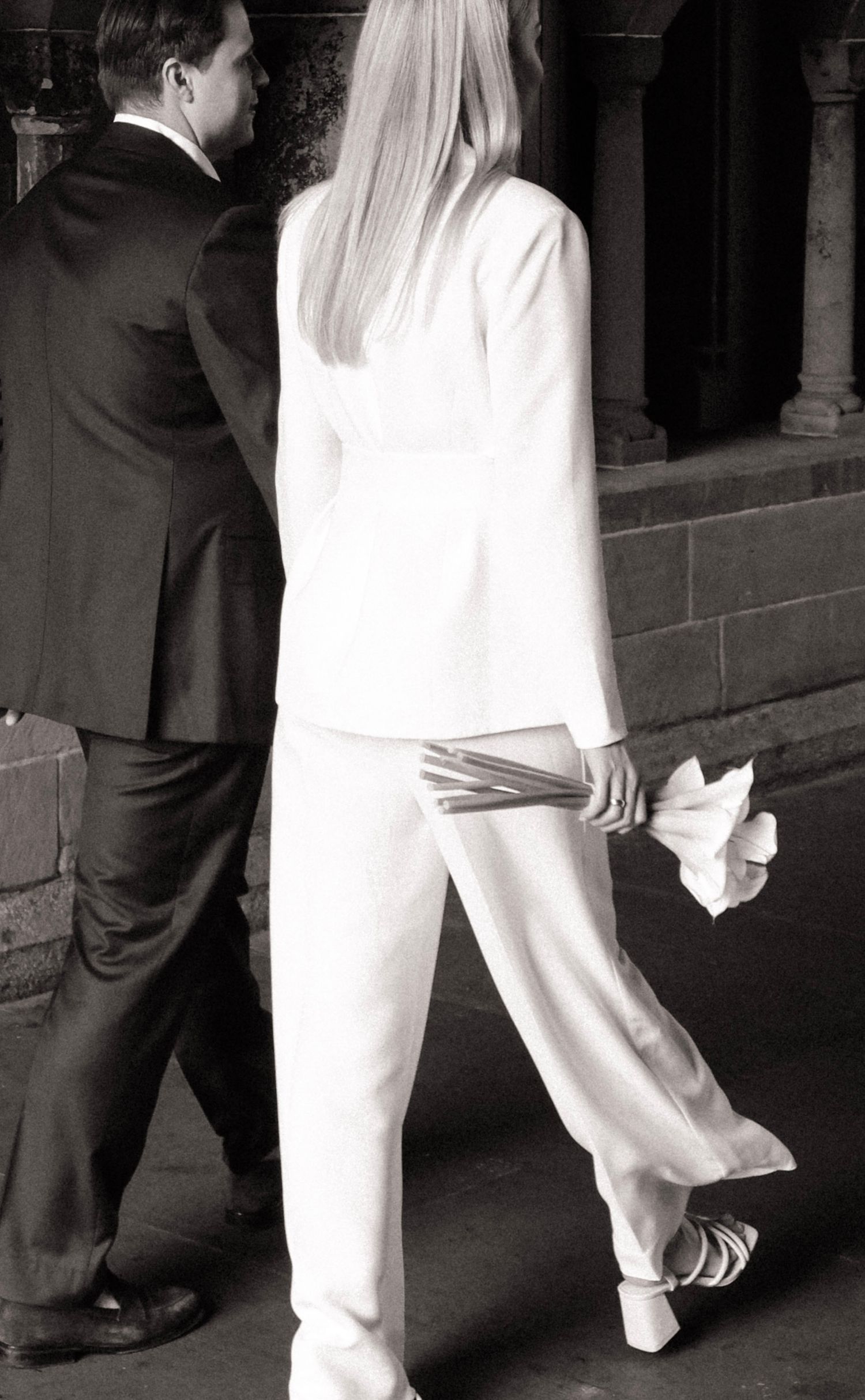 Black and white photo of bride in white pantsuit holding calla lilies walking with suited groom.