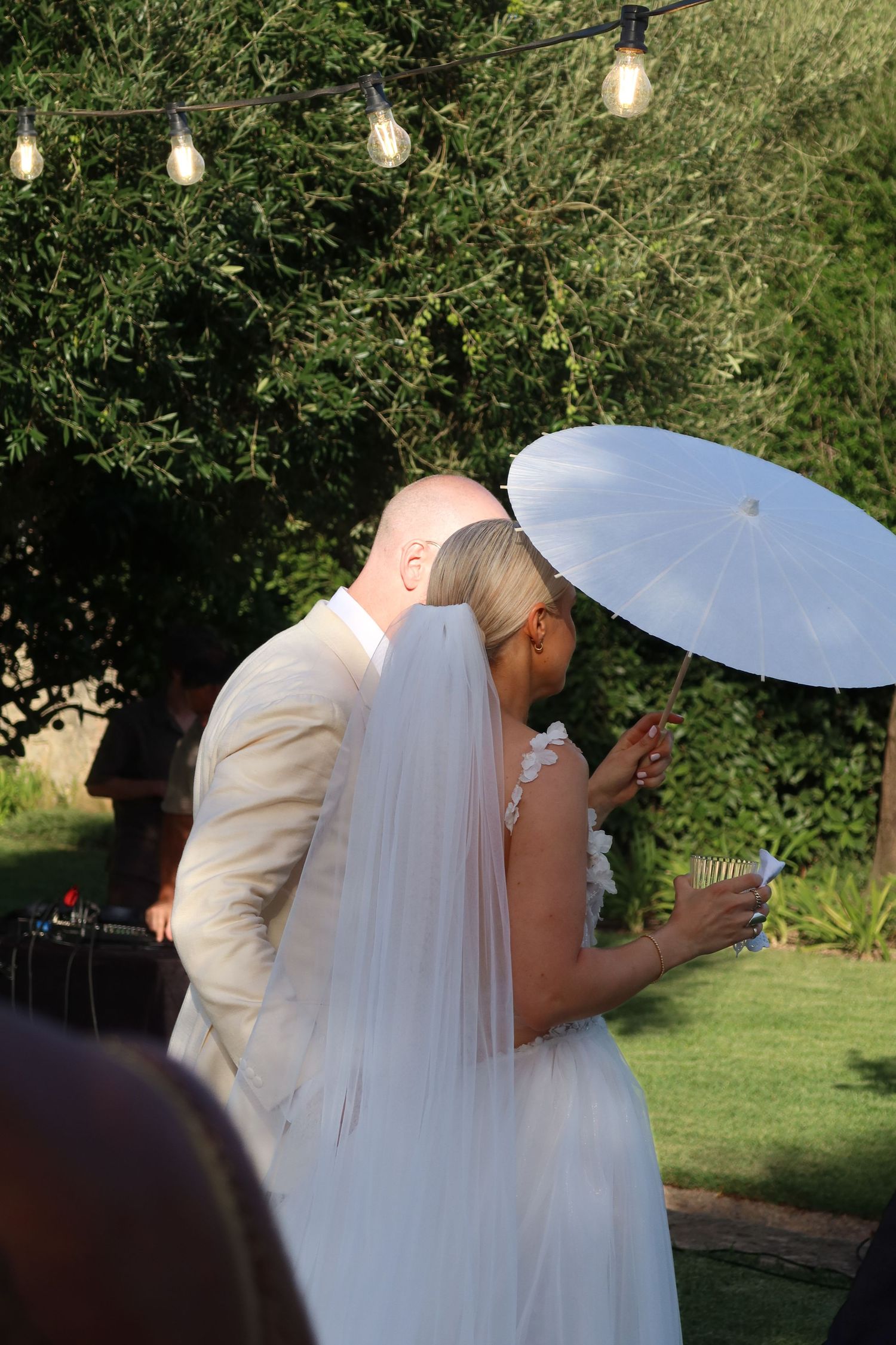 Bride and groom sharing a kiss outdoors under string lights, bride holding a white parasol and veil.