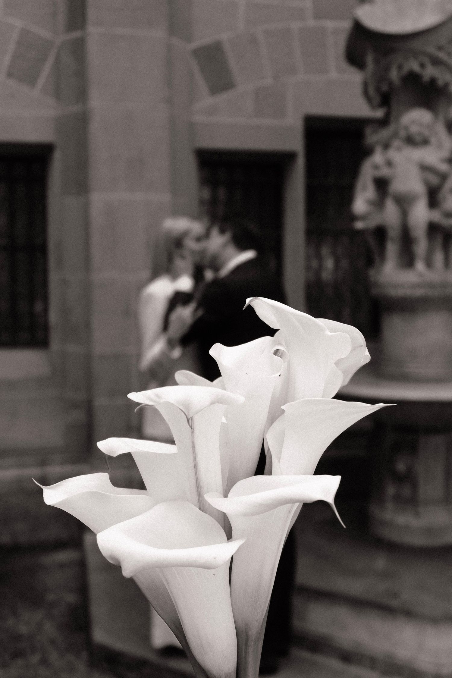 Black and white artistic shot of white calla lily bouquet with kissing couple in background.