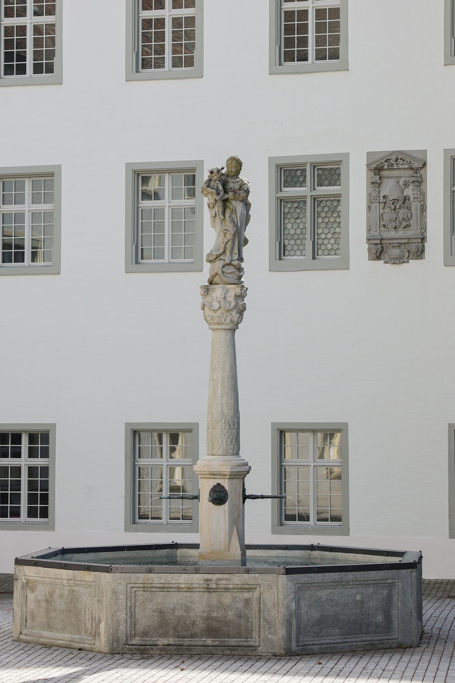 Stone baroque fountain with sculpture on tall column in courtyard of white classical building.