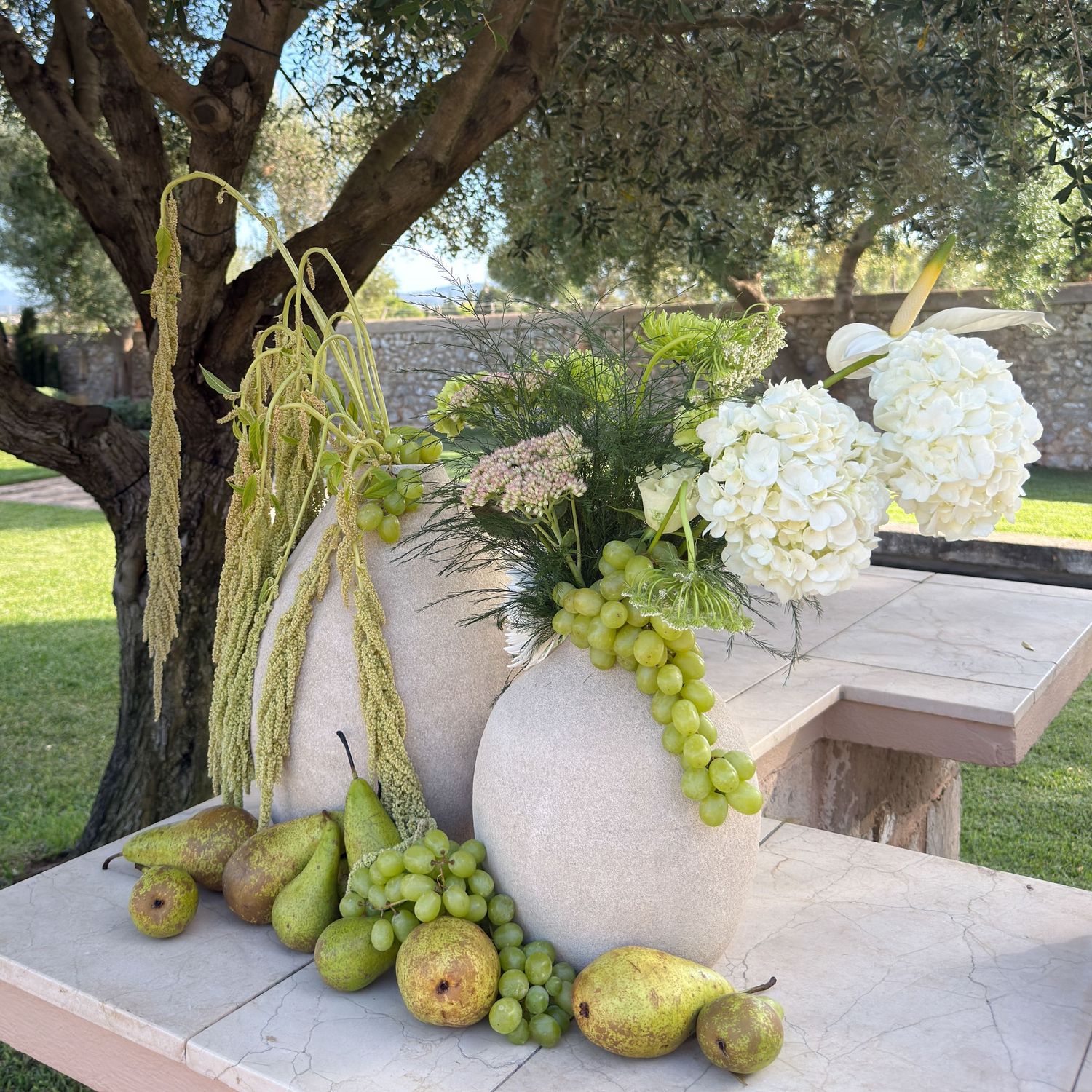 Elegant outdoor wedding floral display featuring white hydrangeas, green grapes, and pears under an olive tree.