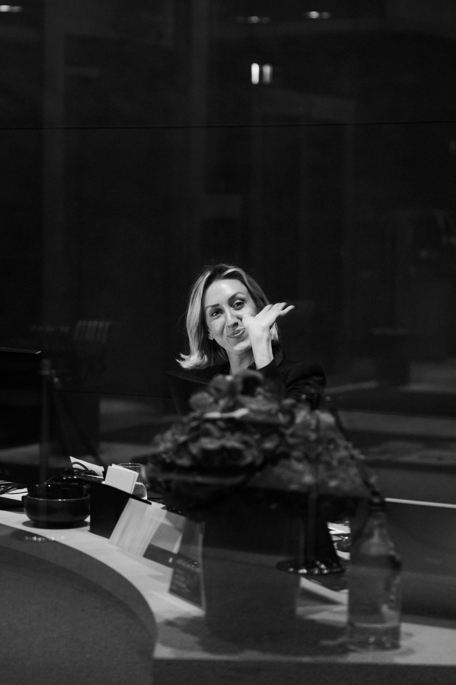 Black and white portrait of a blonde woman gesturing expressively while seated at a dining table indoors.