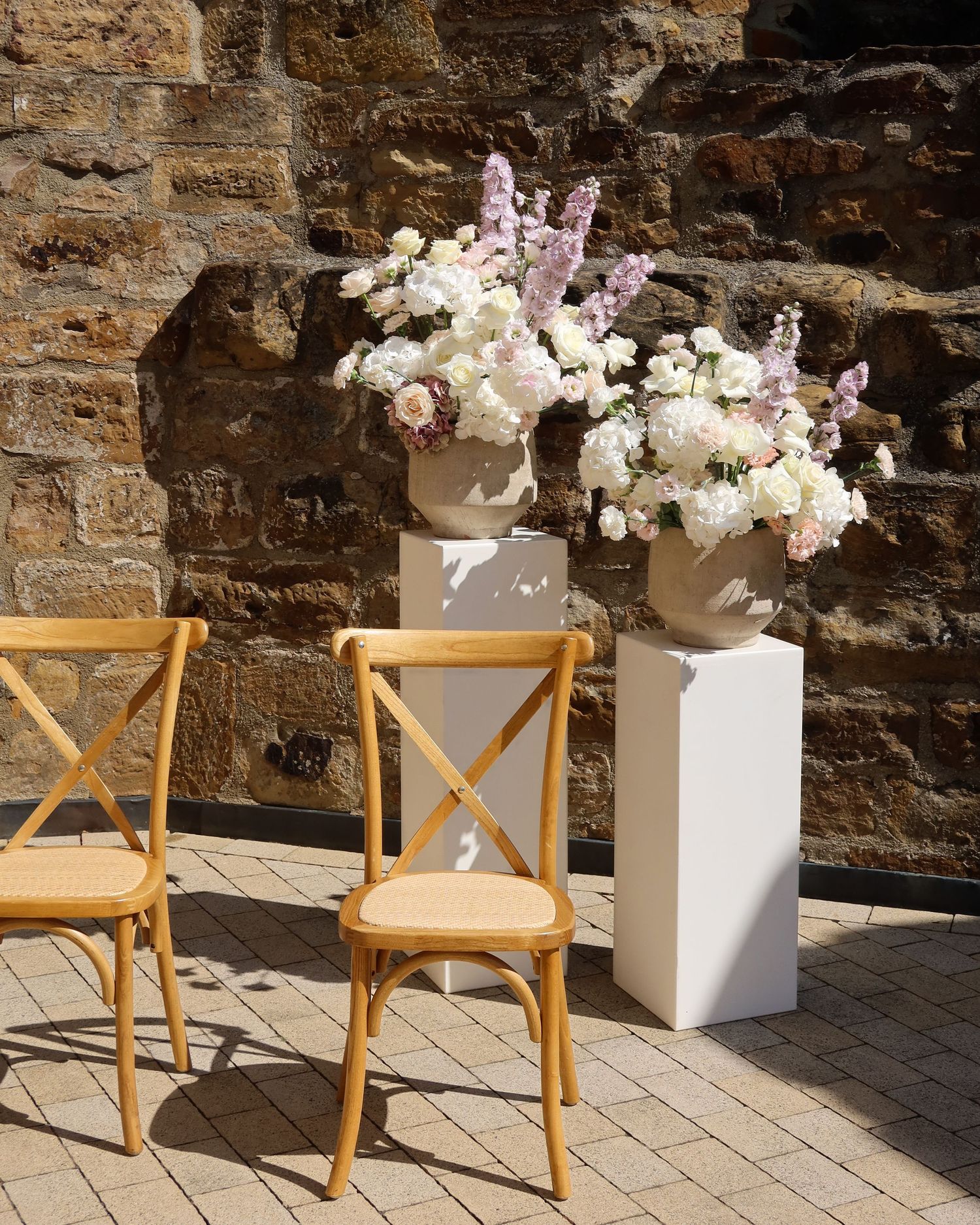 Two large white vases with lush white and lavender floral arrangements on pedestals against a stone wall.