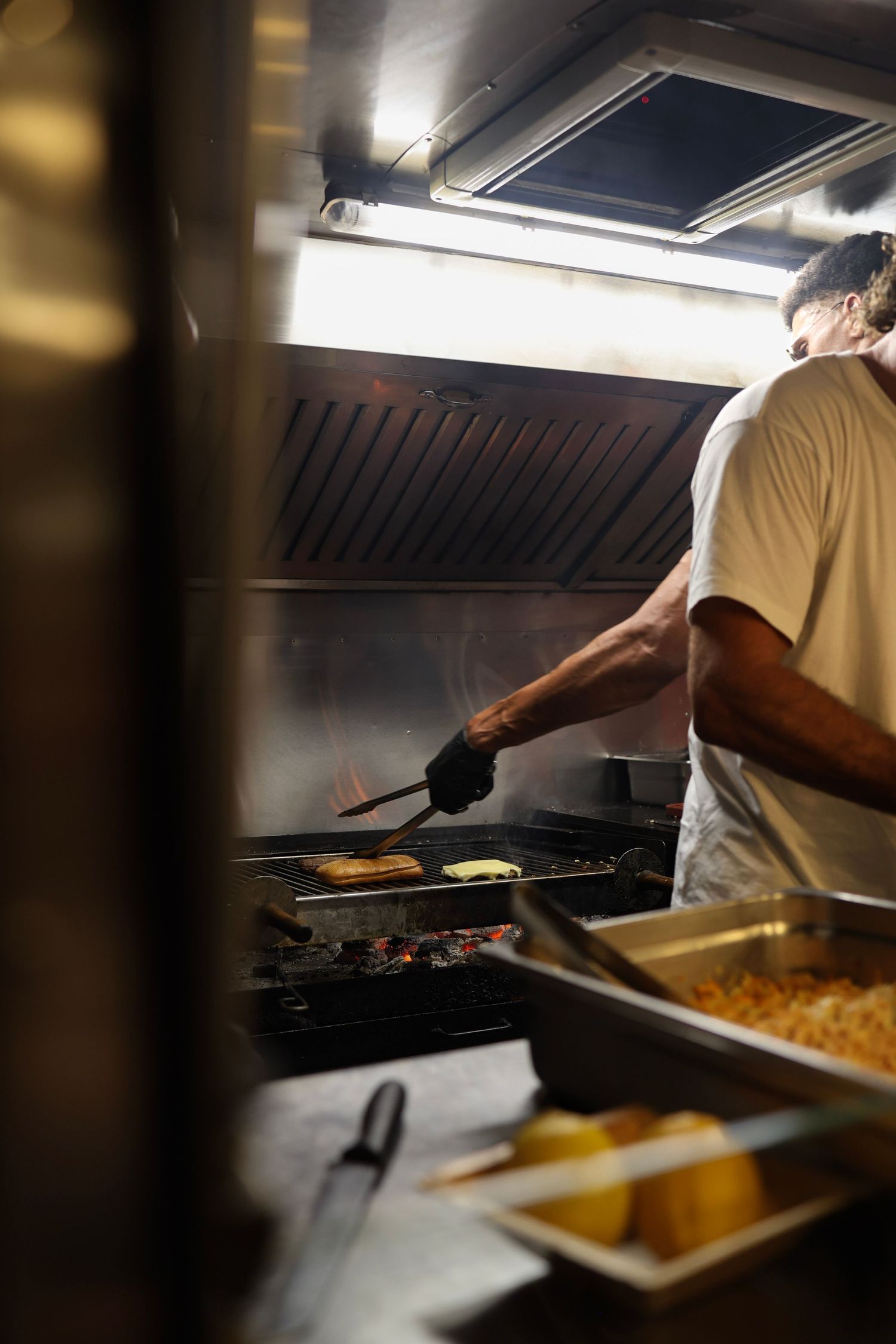 Chef grilling food on a commercial kitchen grill with tongs, smoke rising under a ventilation hood.