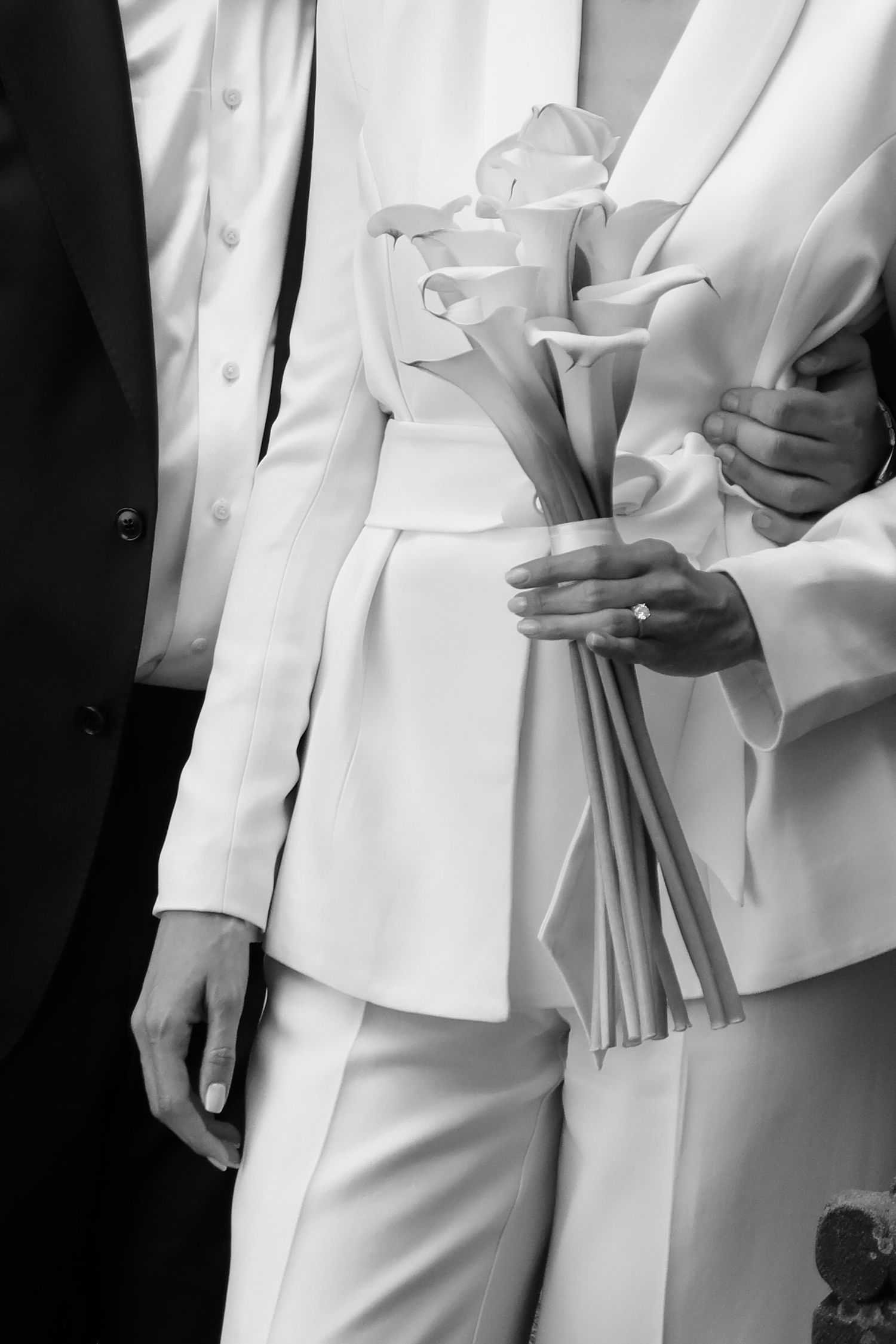 Bride in white suit holding elegant calla lily bouquet with engagement ring visible, black and white wedding photo.