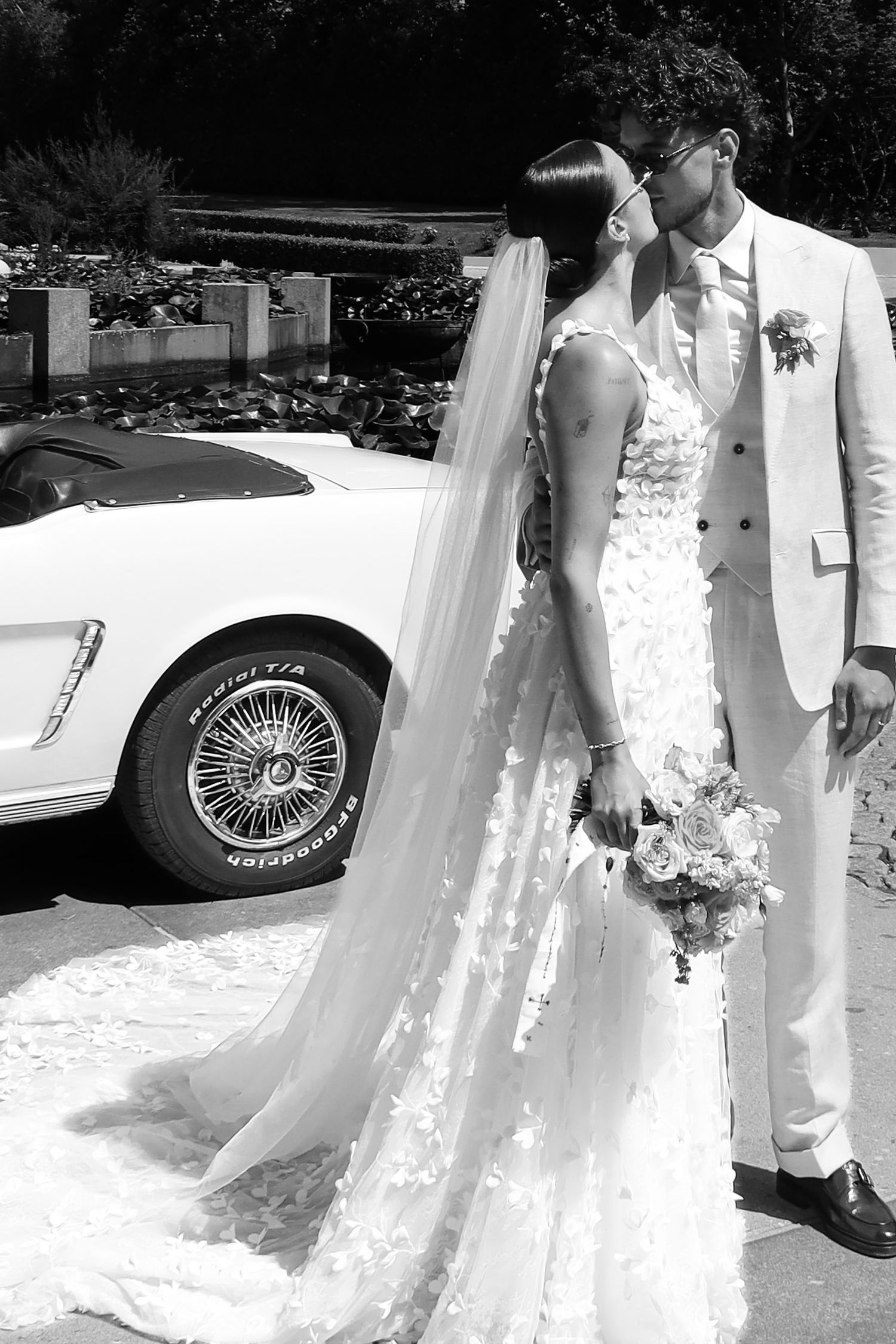 Bride and groom sharing a kiss beside a vintage white sports car, black and white wedding portrait.