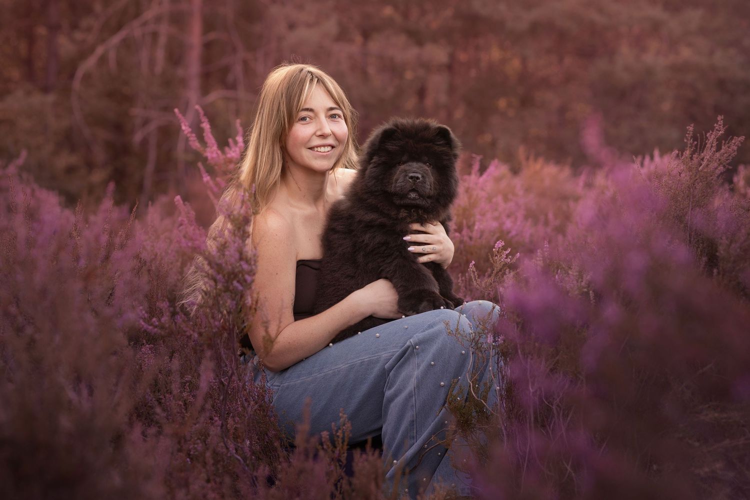 Smiling blonde woman holding a fluffy black Chow Chow dog in a field of purple heather flowers.