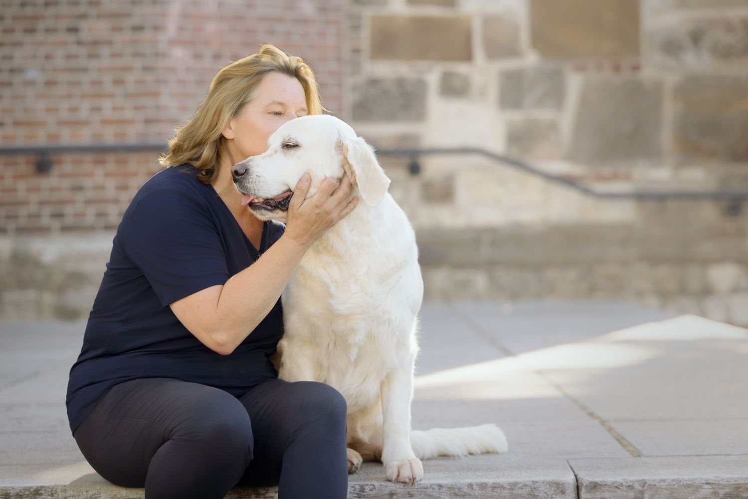 A blonde woman kneels on stone steps hugging a large white Golden Retriever near a brick wall.