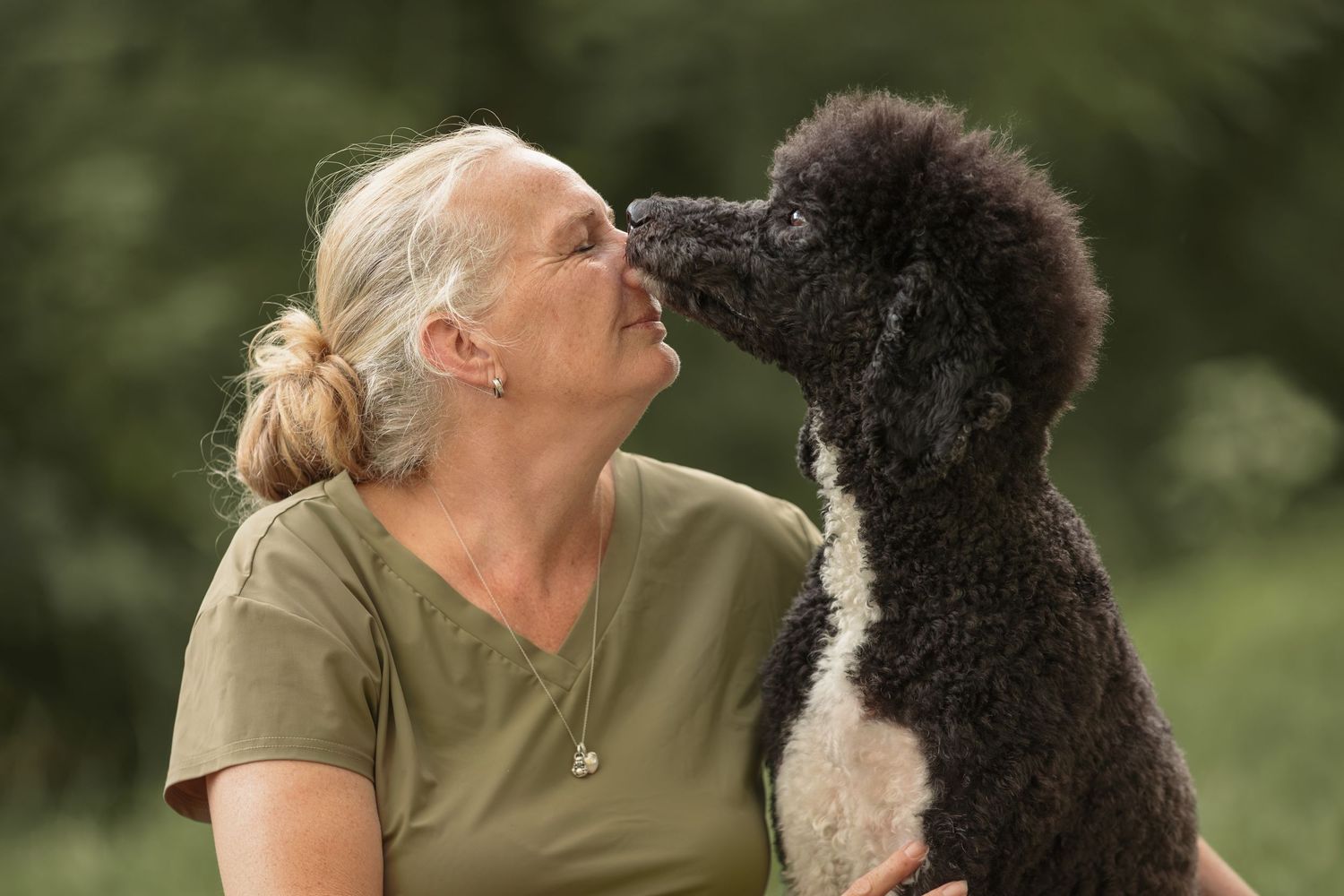 An older woman shares a tender nose-to-nose moment with a black and white Standard Poodle outdoors.