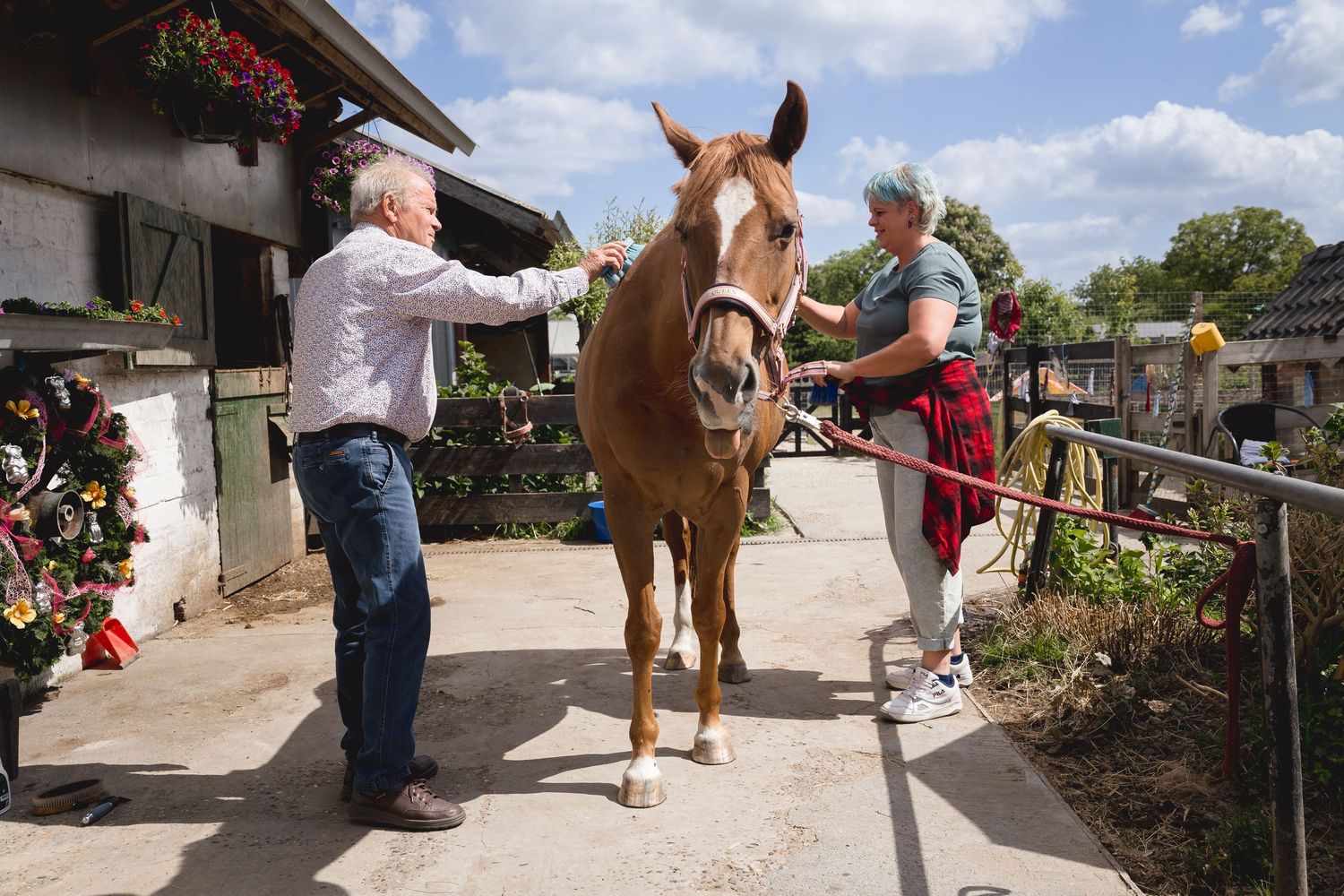 Two people grooming a chestnut horse outside a stable on a sunny day.