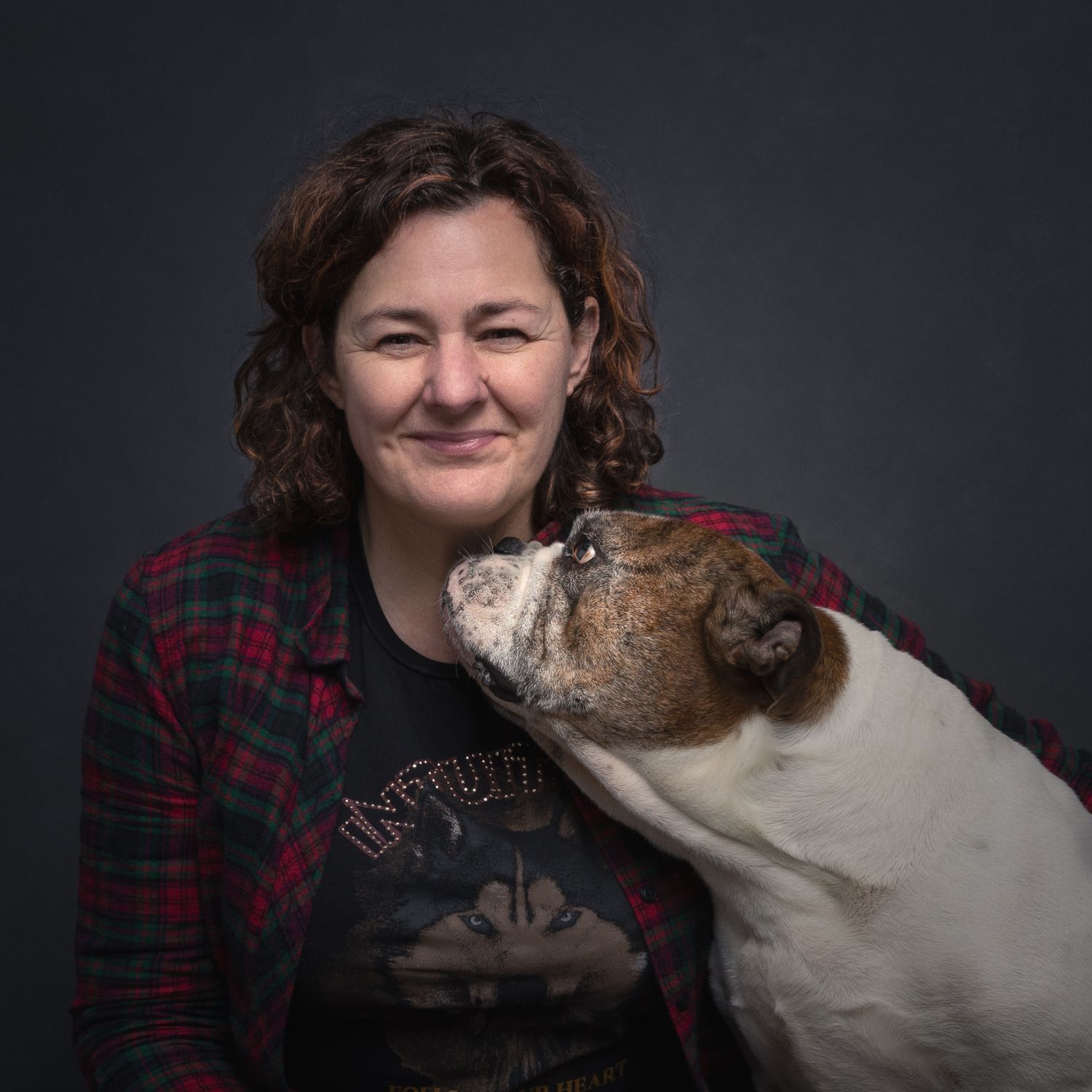 A smiling woman with curly hair is affectionately licked by a brown and white bulldog in a studio.